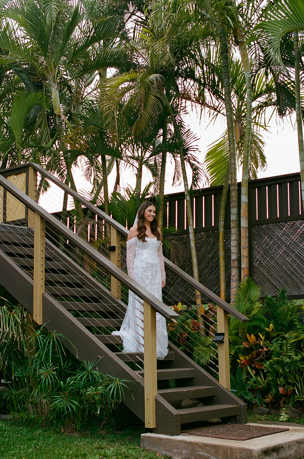 Bride in a beaded lace wedding gown descending outdoor stairs surrounded by tropical Hawaii greenery
