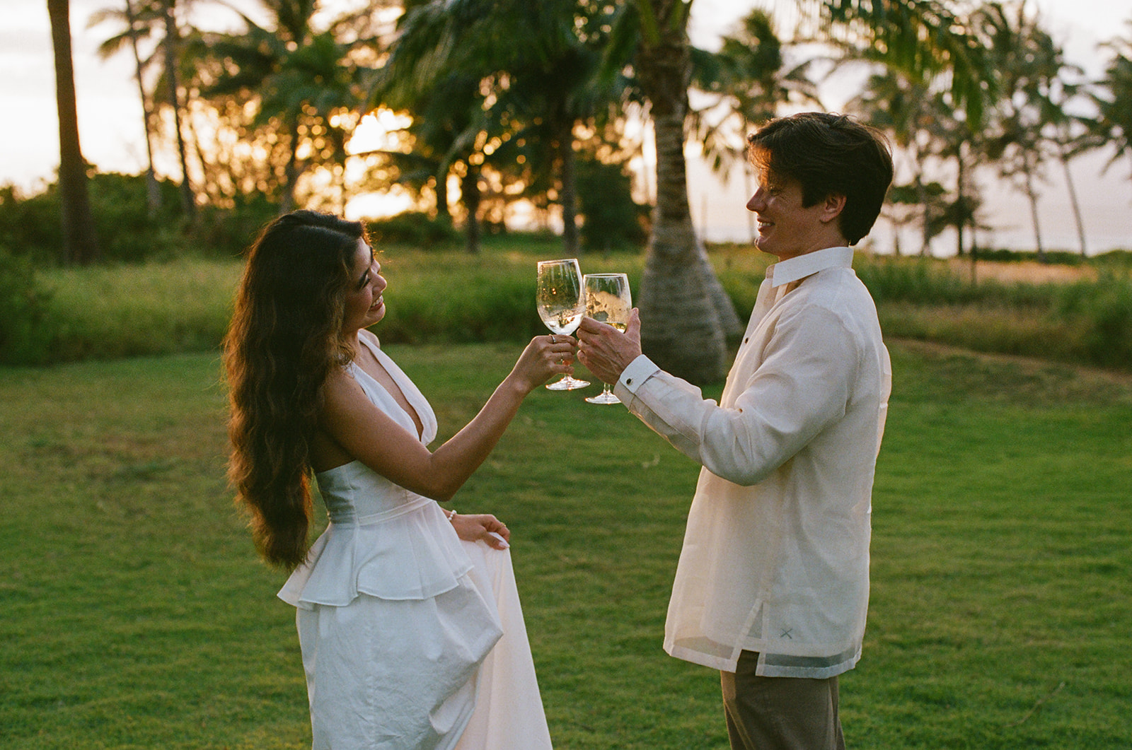 Bride and groom toasting with wine glasses on the lawn at golden hour

