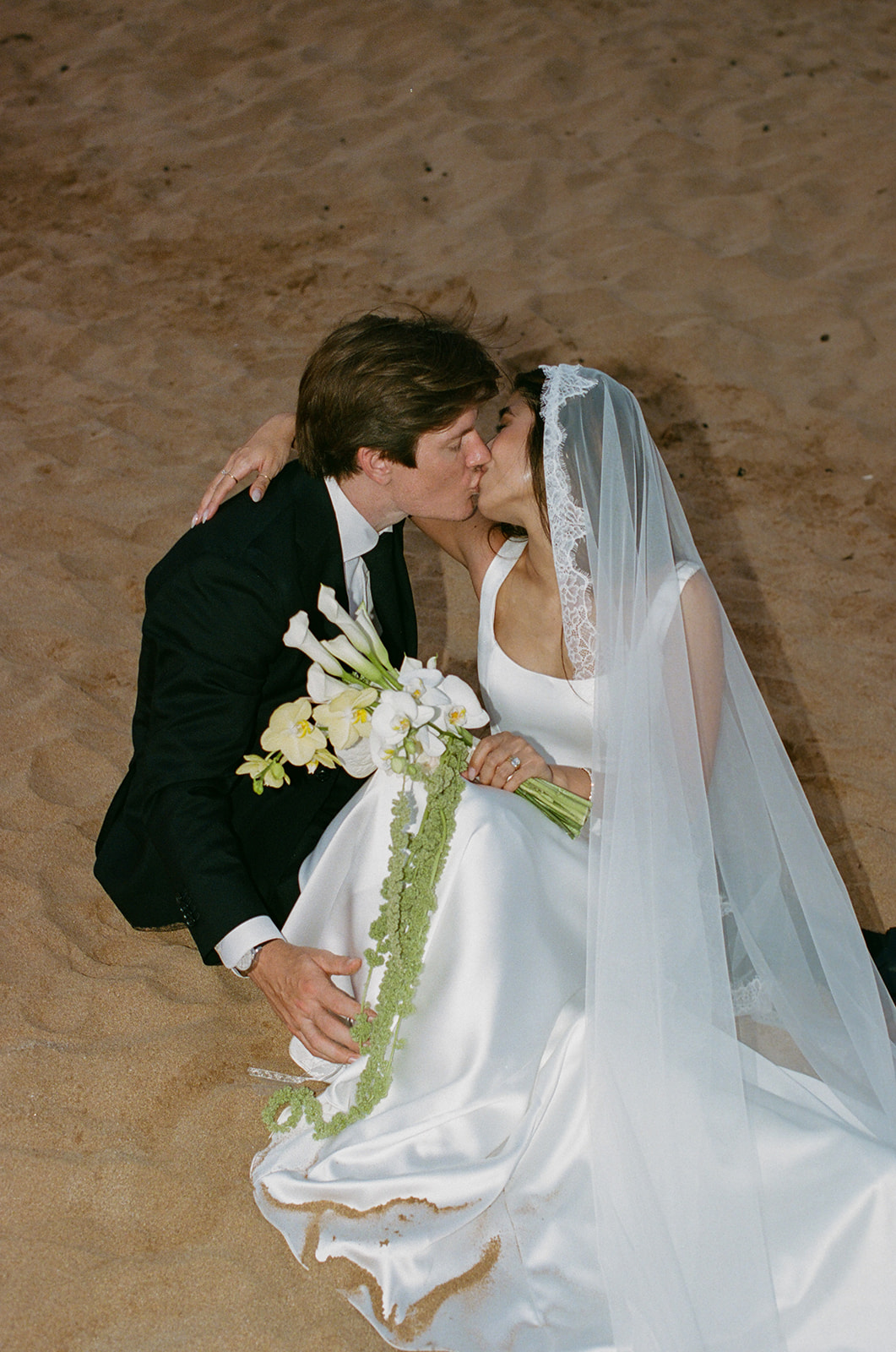 Bride and groom kissing while seated together on the sand at dusk
