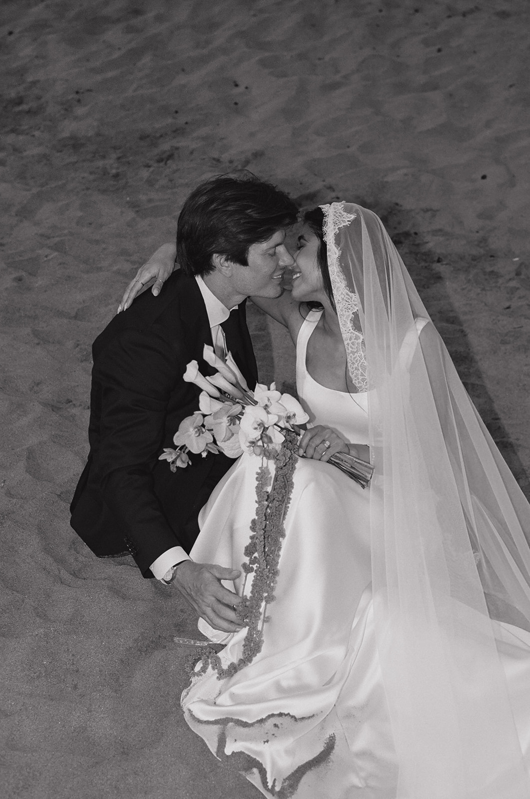 Black and white close-up of the bride and groom kissing while seated on the sand
