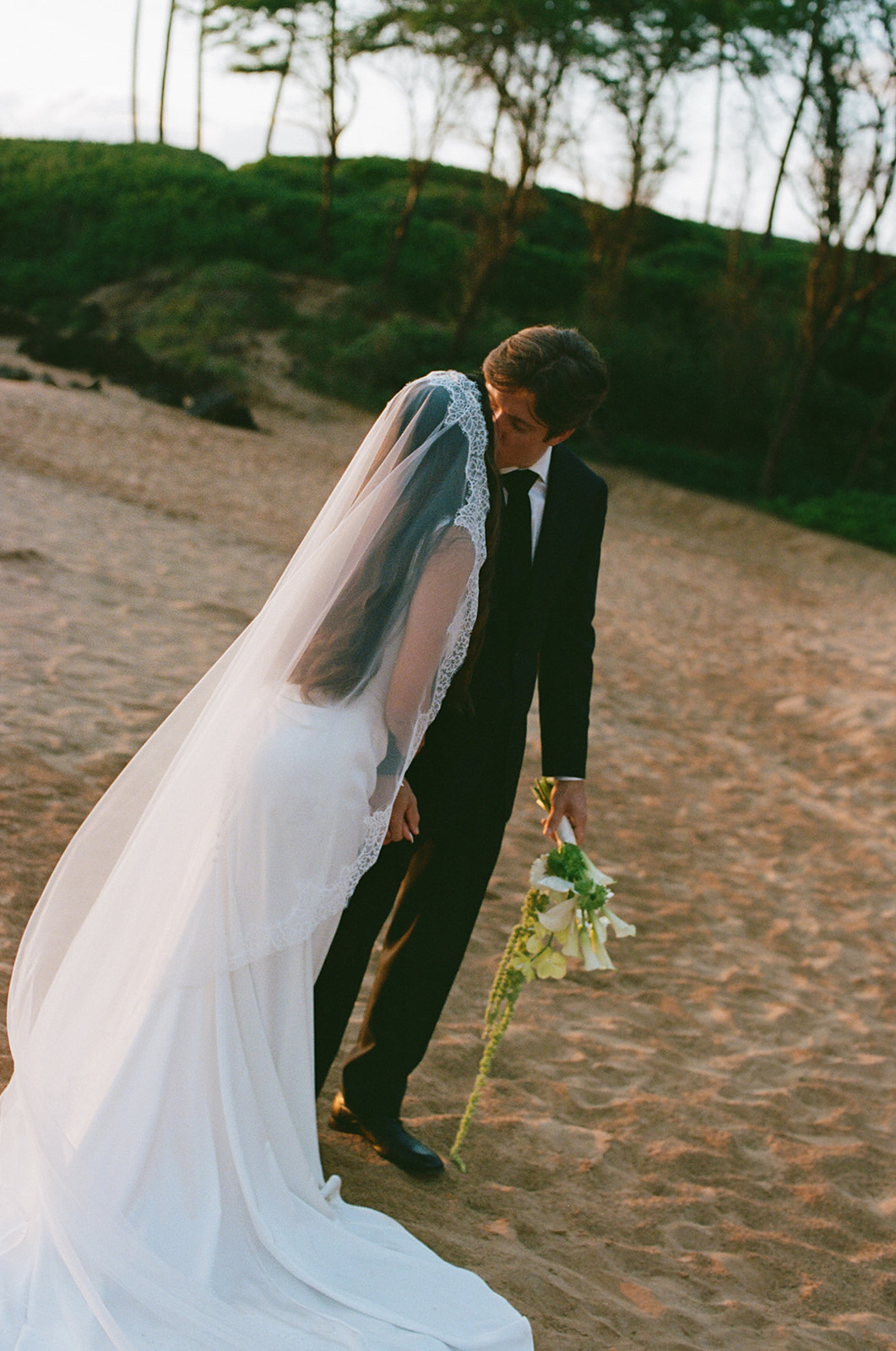 Bride and groom sharing a kiss on the beach as her veil drapes over his shoulder
