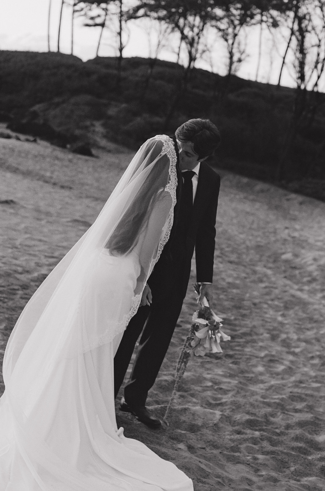 Bride and groom sharing a kiss on the beach in black and white, her veil draped over his shoulder
