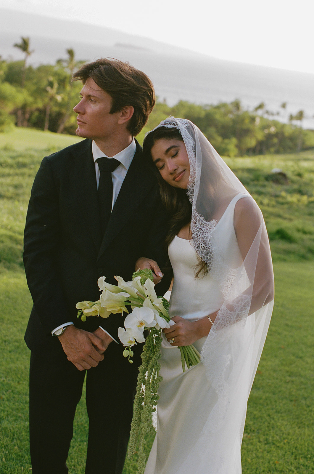 Groom resting his head close to his bride as she leans into him, bouquet in hand
