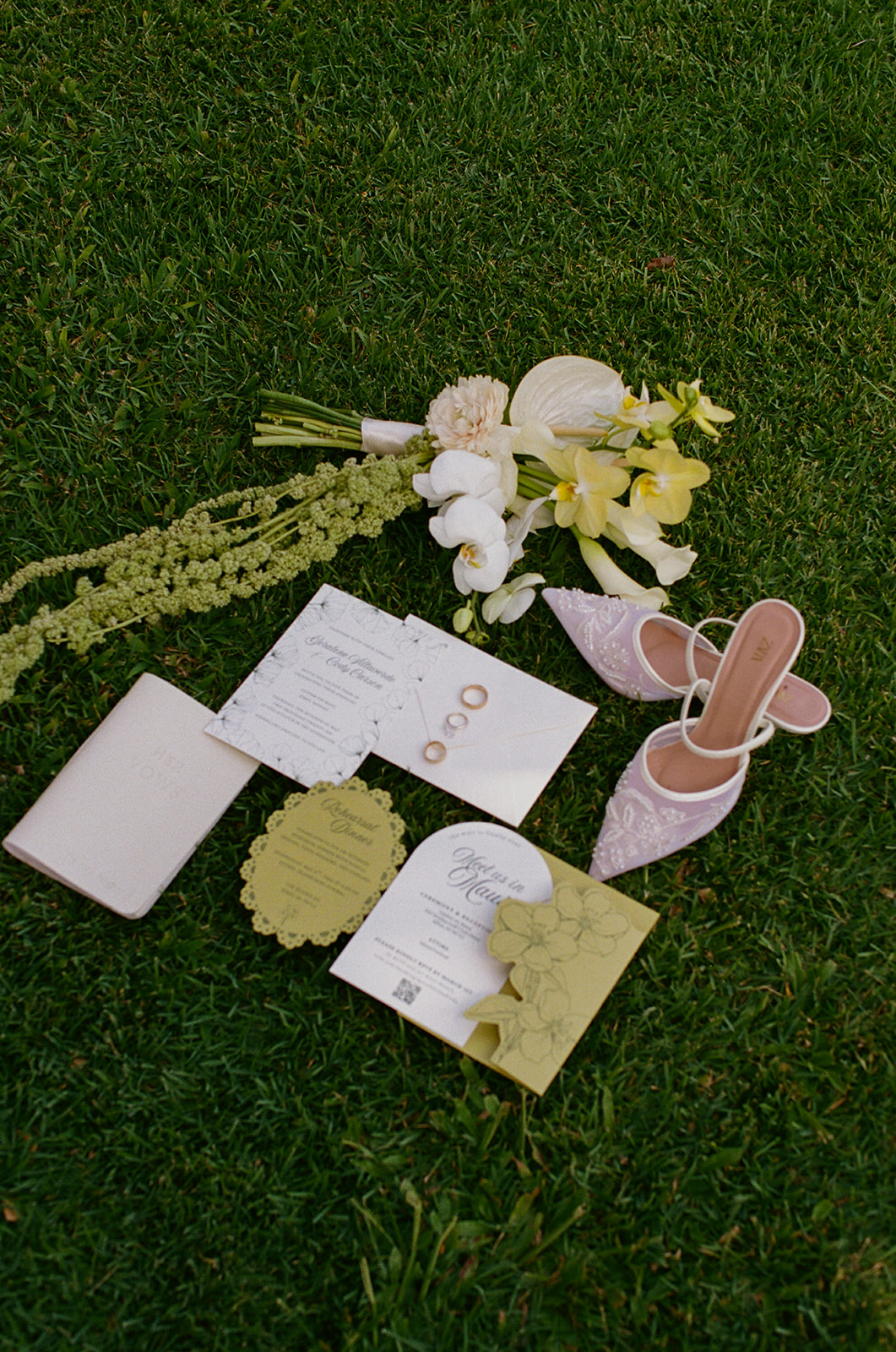 Wedding film photography flat lay of invitations, rings, lavender heels, and a yellow-white bouquet on the grass
