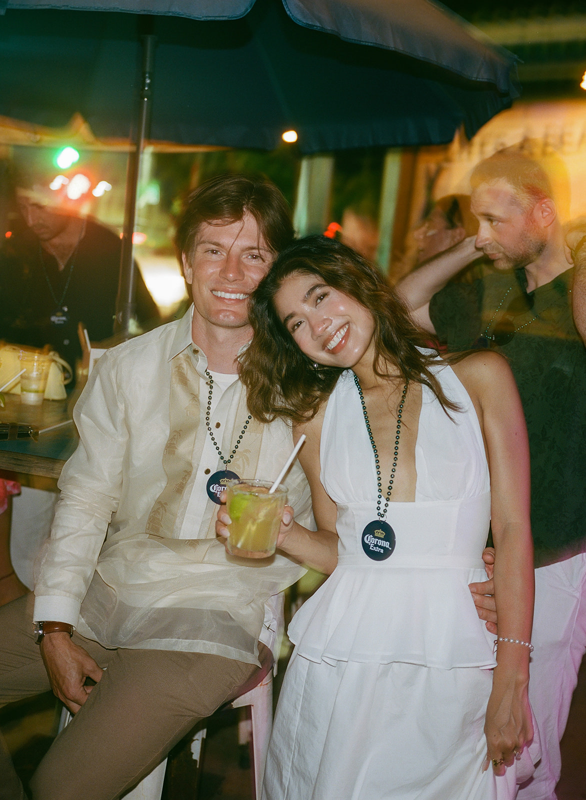 Wedding film photography of the newlyweds smiling together at a beachside bar with drink leis
