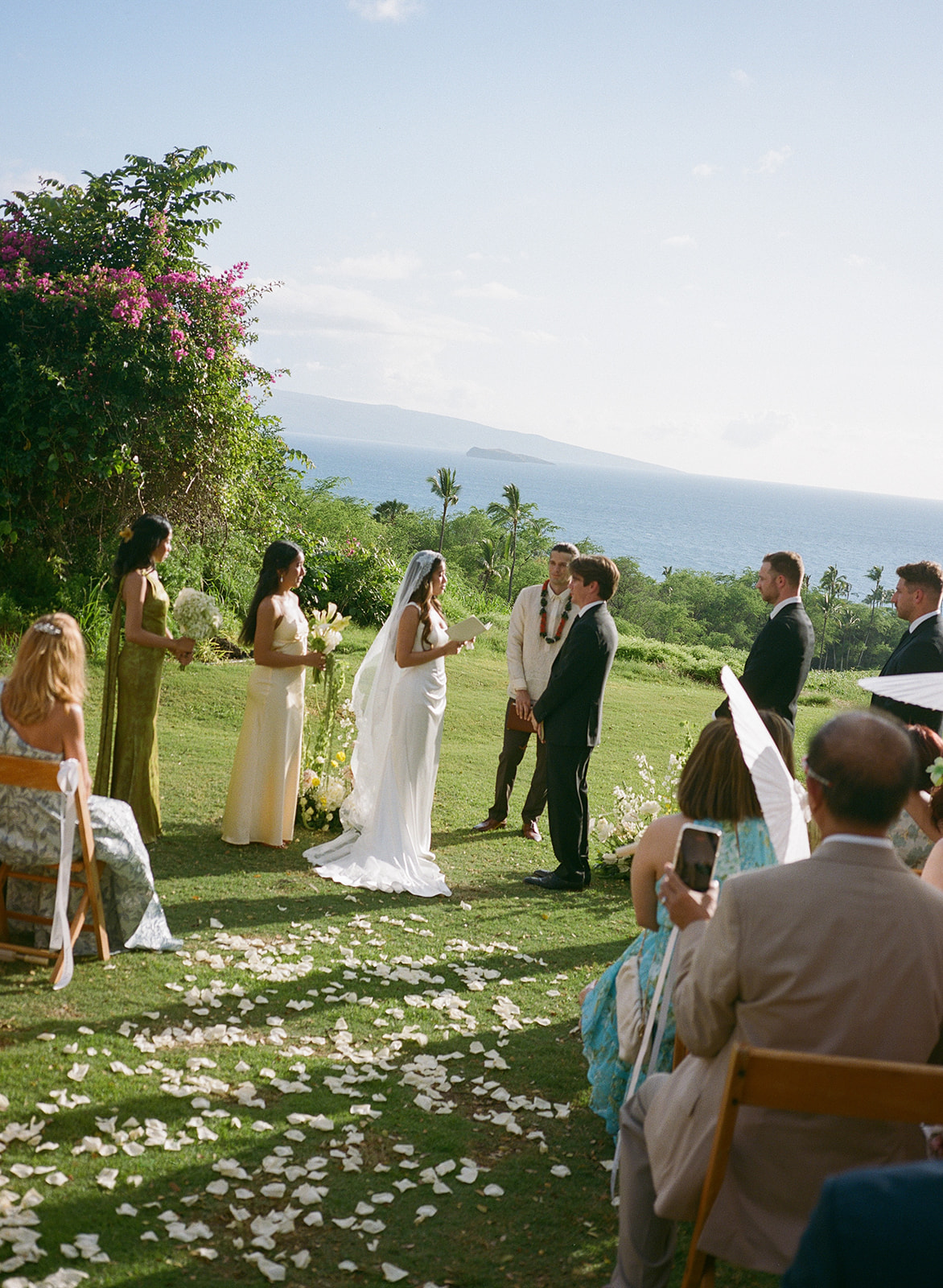 Bride and groom standing before their officiant during an oceanview ceremony with bridesmaids and guests

