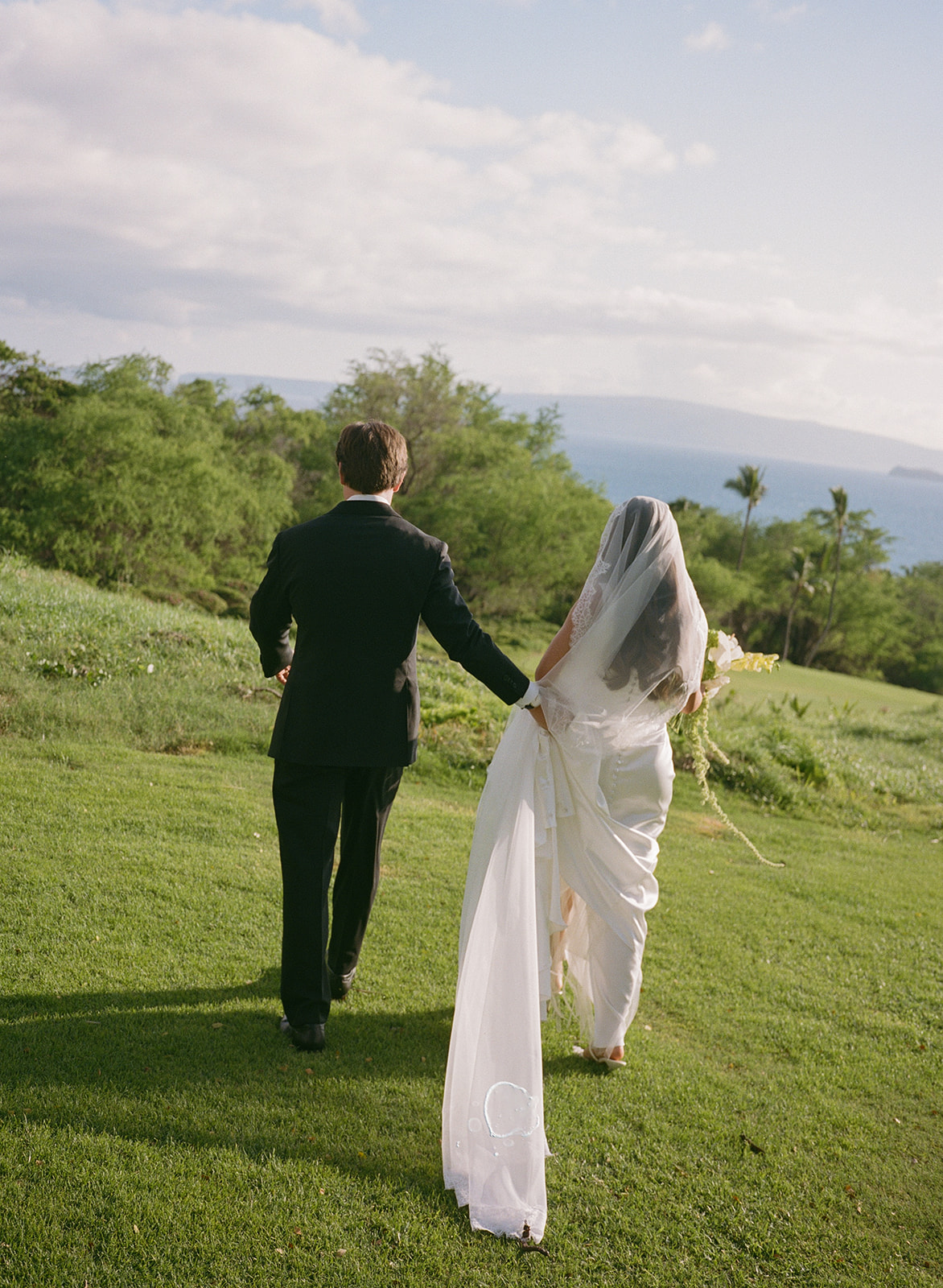 Bride and groom walking hand in hand across a grassy bluff overlooking the ocean

