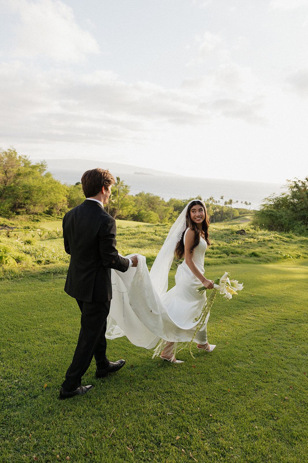 Bride glancing back with a smile as the groom carries her dress train across the lawn

