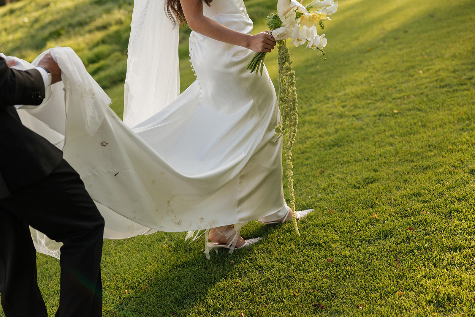 Wedding film photography moment of the groom lifting the bride's train as she walks across the lawn
