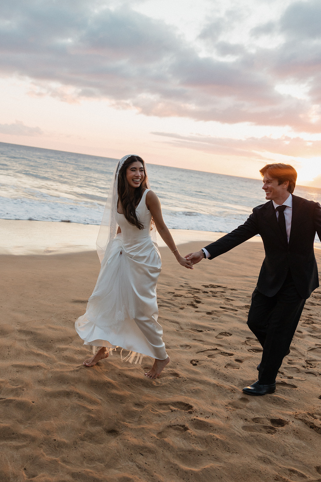 Bride laughing as she holds the groom's hand while running along the beach at sunset

