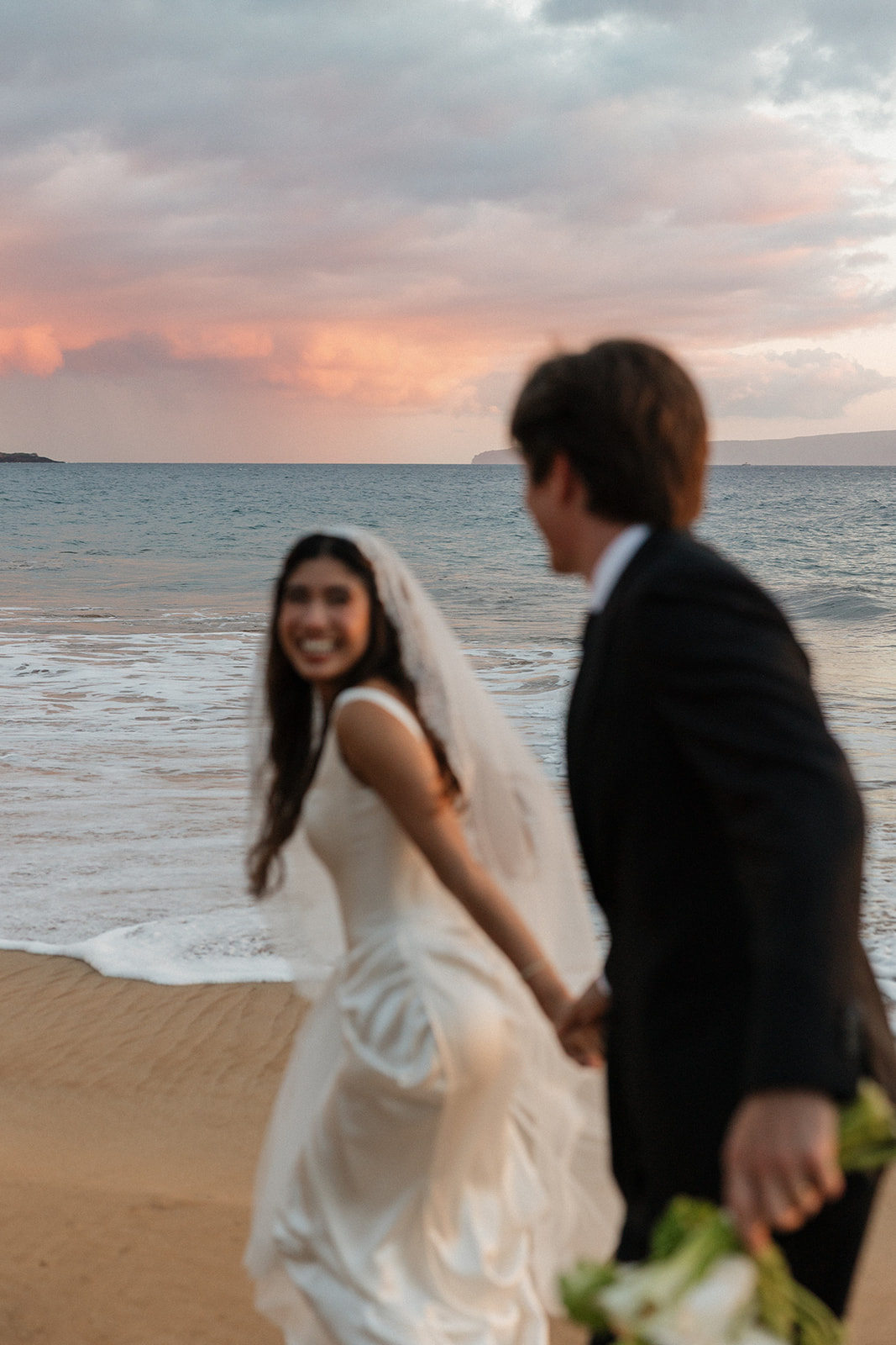 Wedding film photography of the bride laughing over her shoulder as the couple strolls along the shoreline
