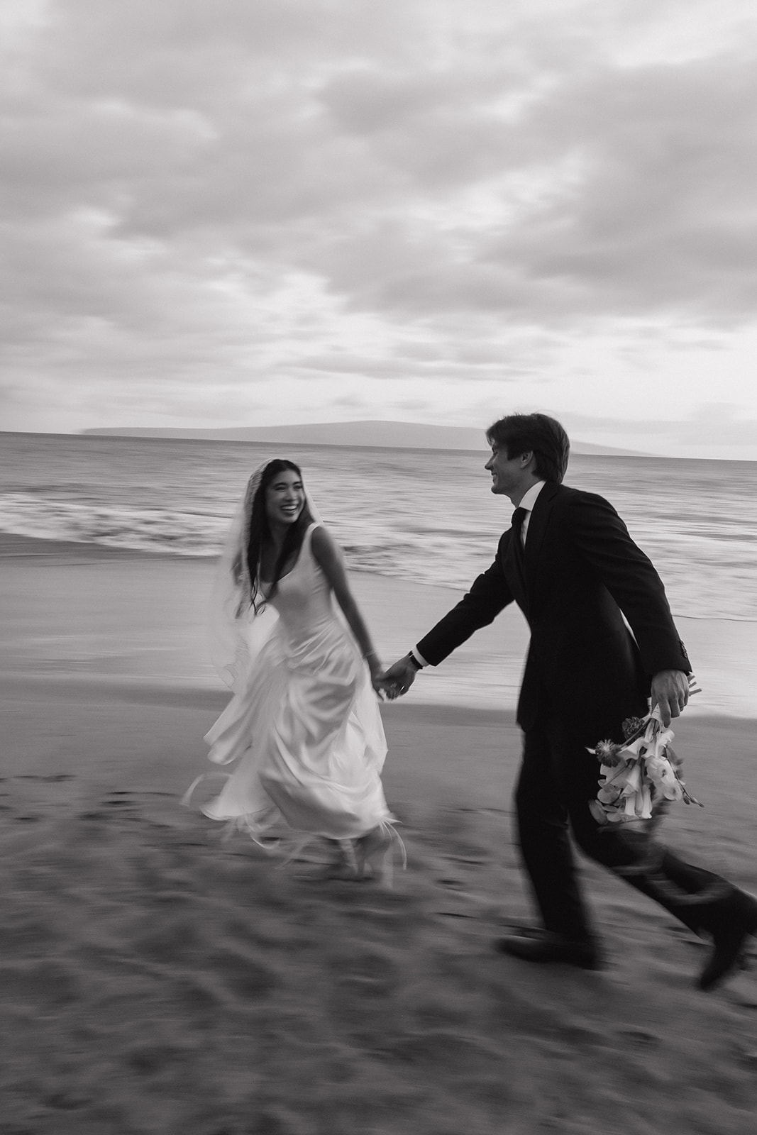 Black and white candid of the bride and groom laughing as they walk hand in hand on the shore
