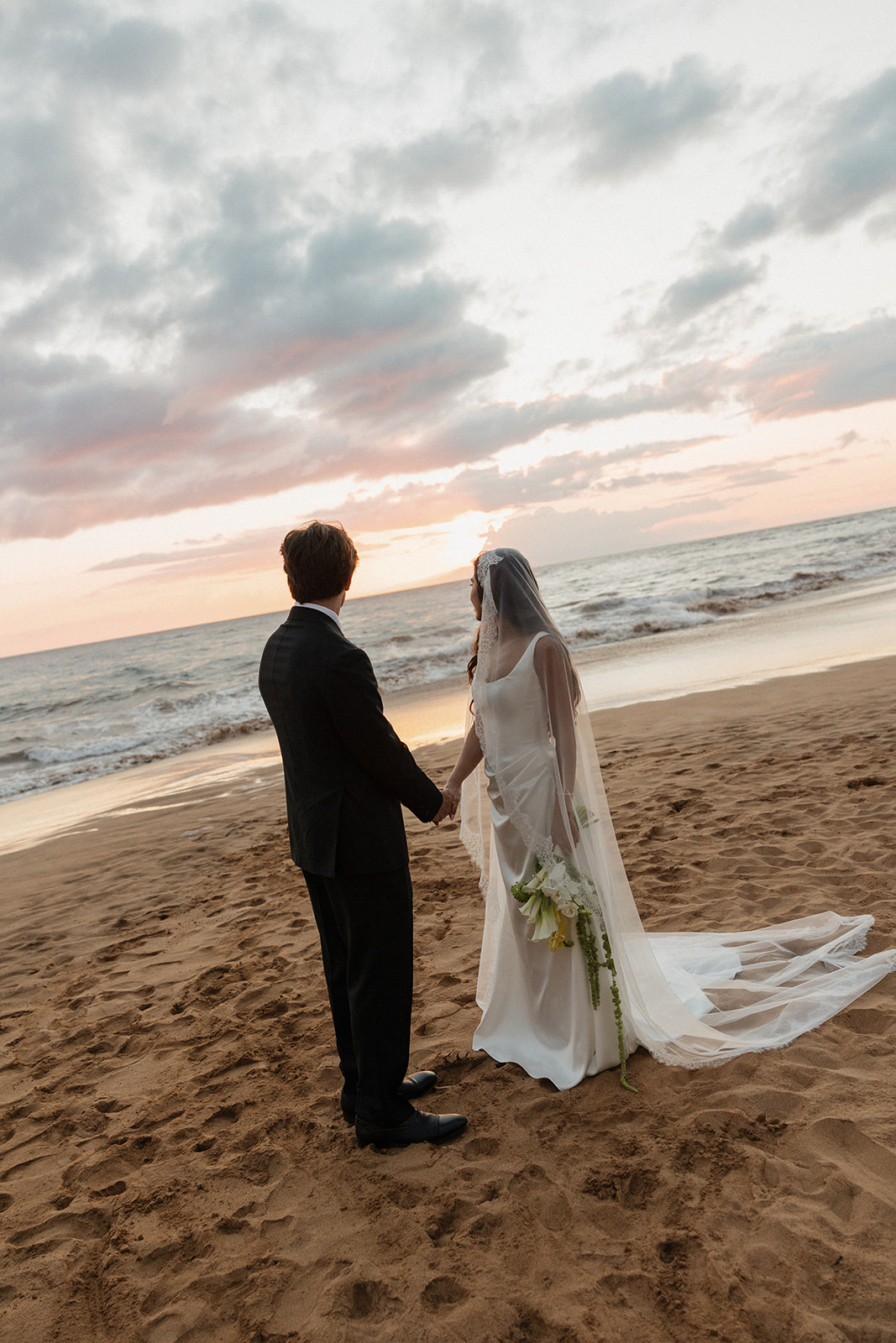 Bride and groom holding hands as they gaze out at the ocean at sunset
