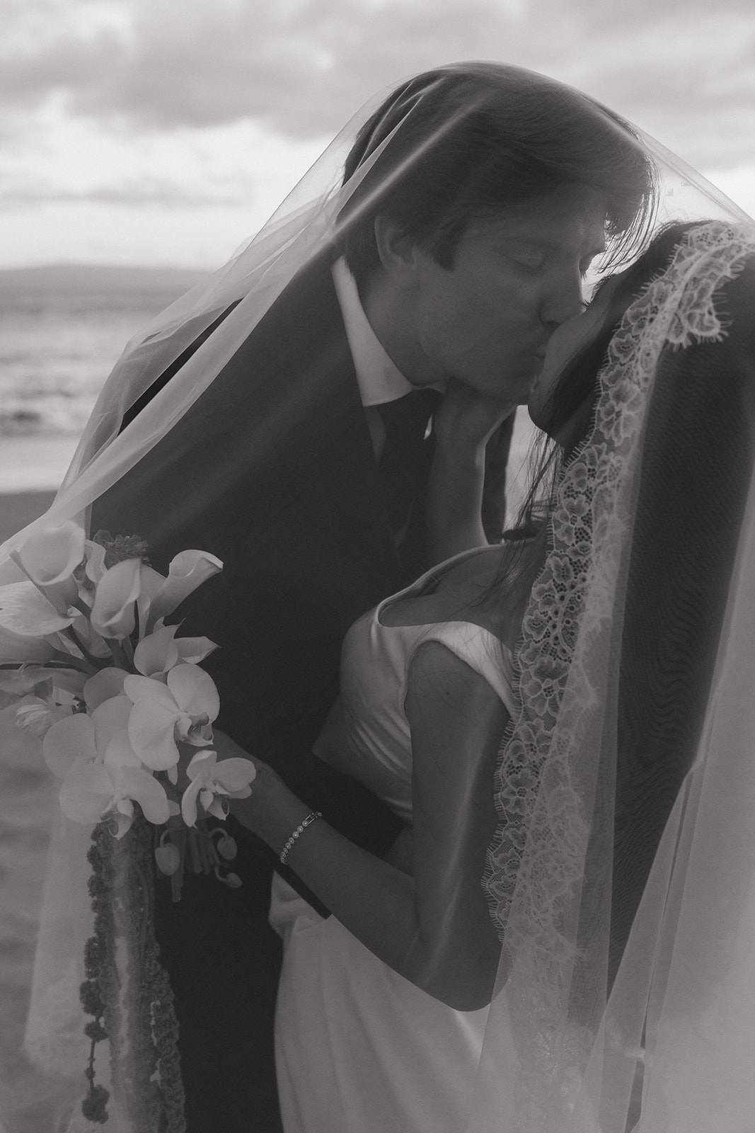 Wedding film photography close-up of the bride and groom kissing under her lace veil at the beach
