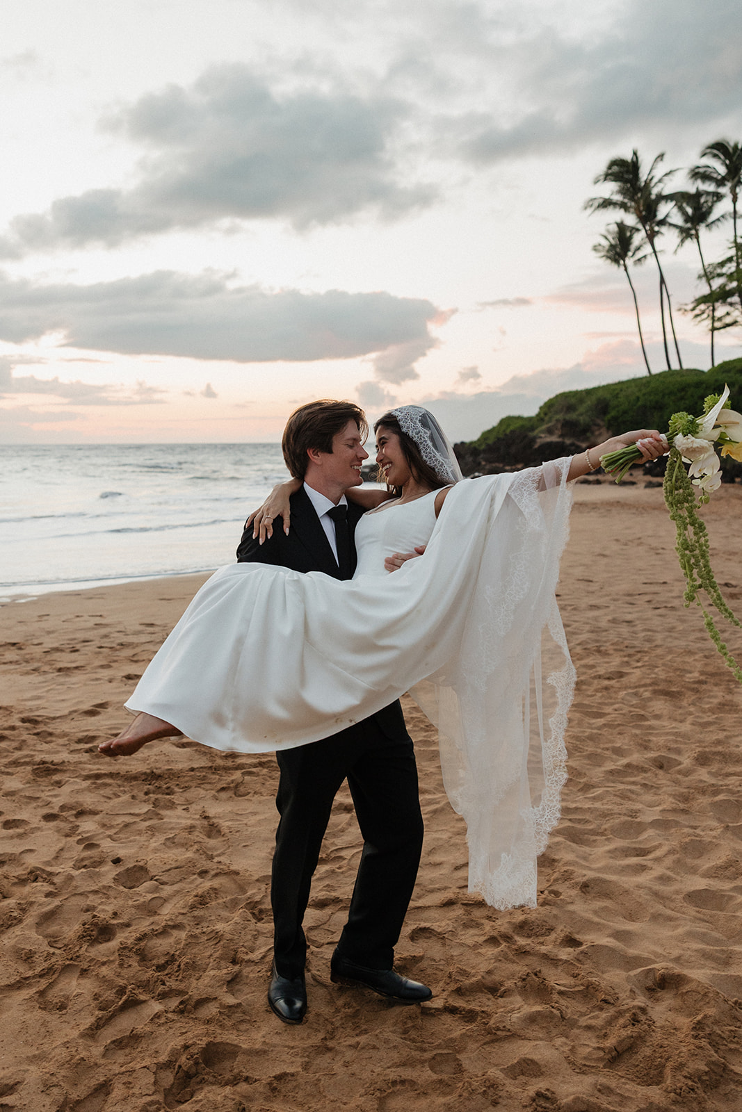 Groom carrying his bride across the beach as she reaches out with her bouquet
