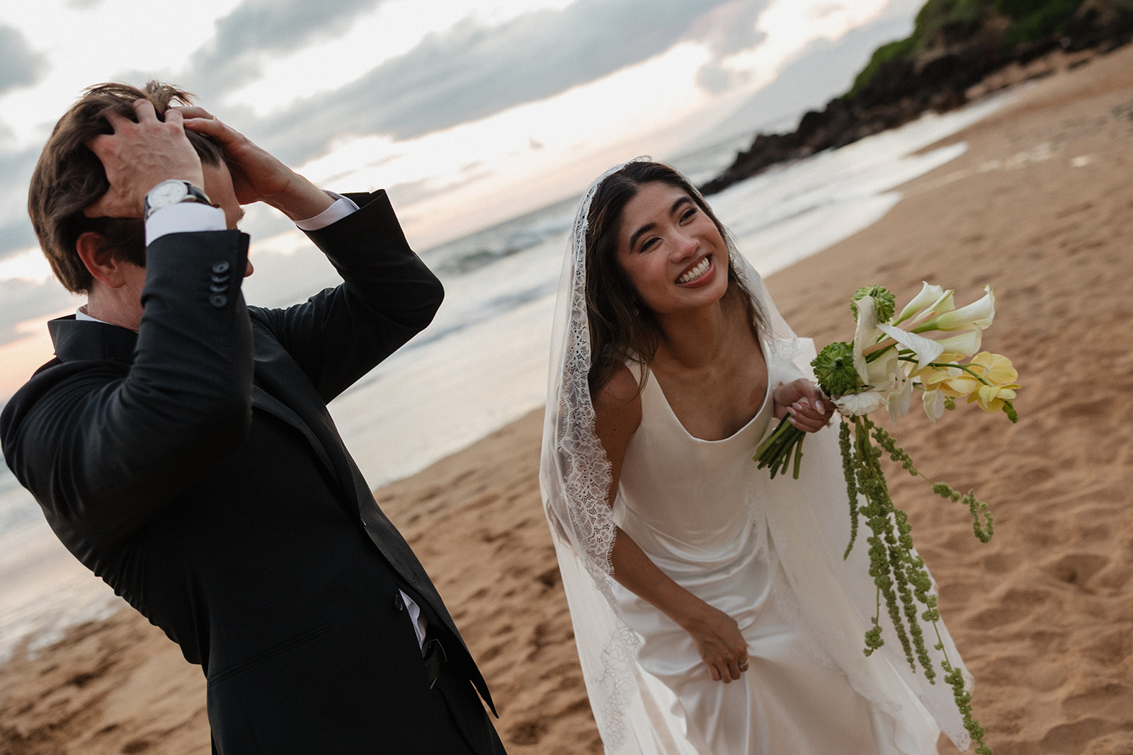Wedding film photography of the groom running his hands through his hair beside his smiling bride on the beach
