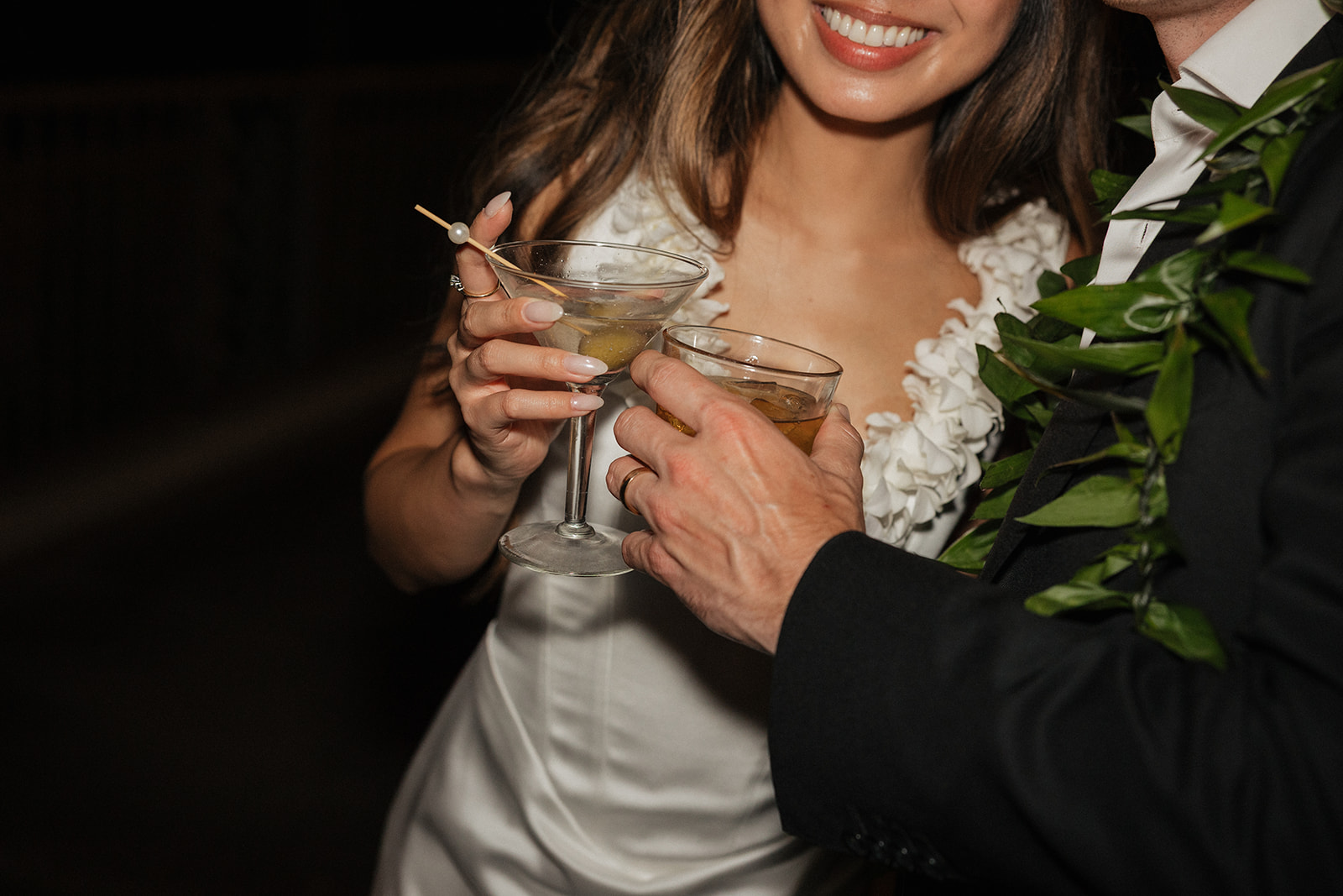 Close-up of the bride and groom clinking cocktail glasses, his lei visible beside her
