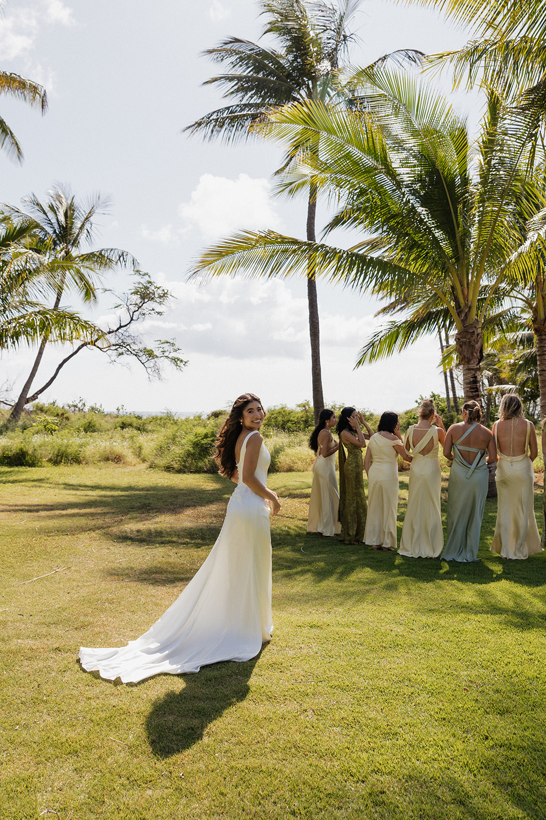 Bride glancing back with a smile as her bridesmaids gather near palm trees
