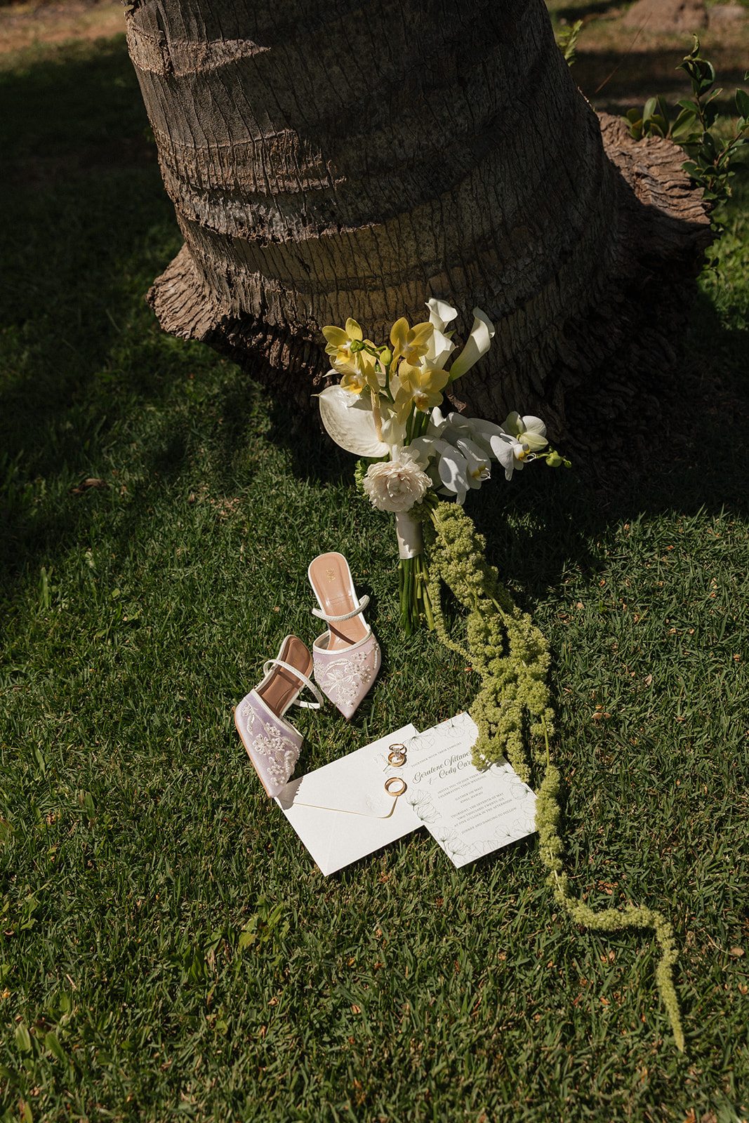 Wedding film photography flat lay of the bridal bouquet, heels, and invitation suite under a palm tree
