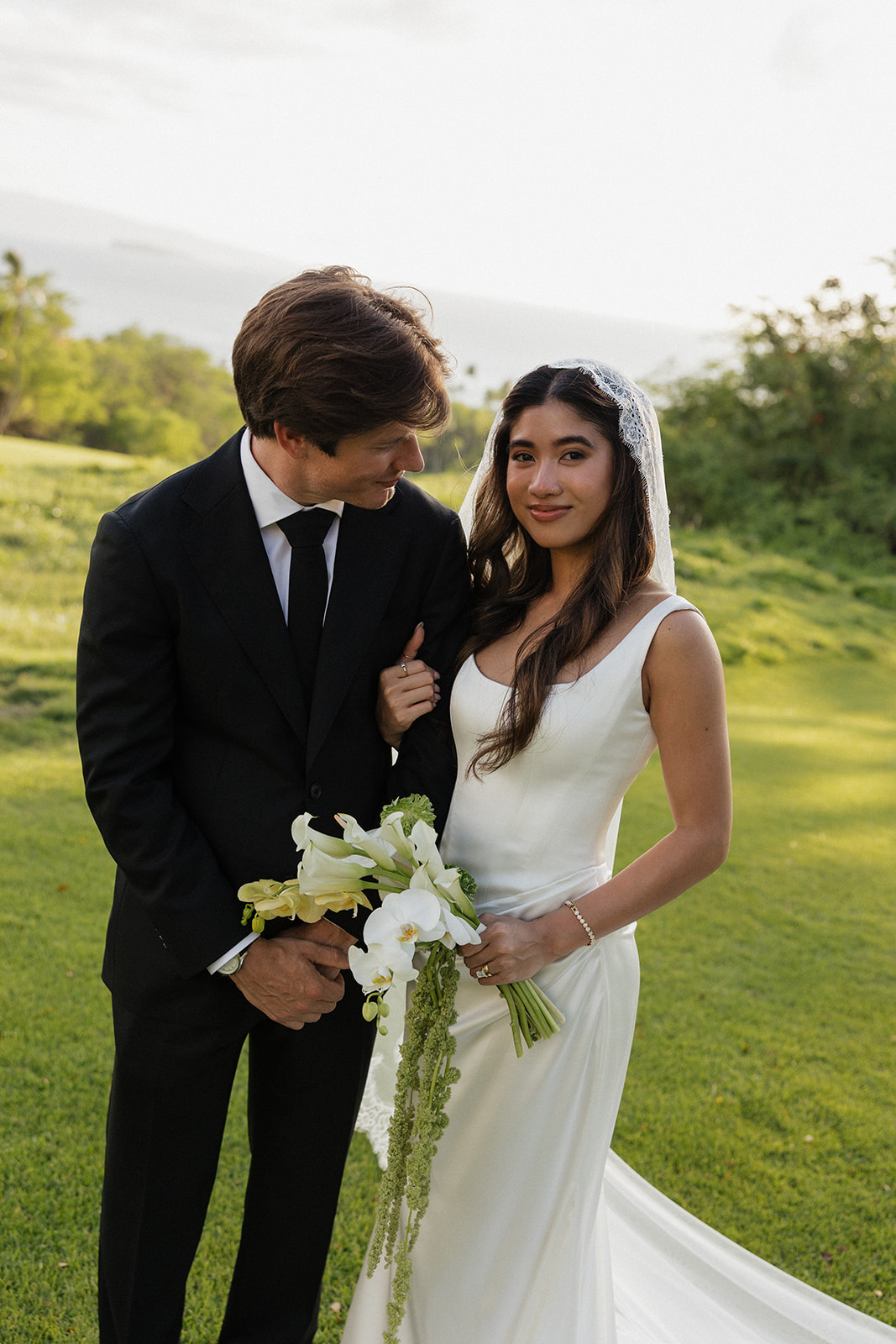 Bride and groom standing close together on the lawn, her veil and bouquet catching the light
