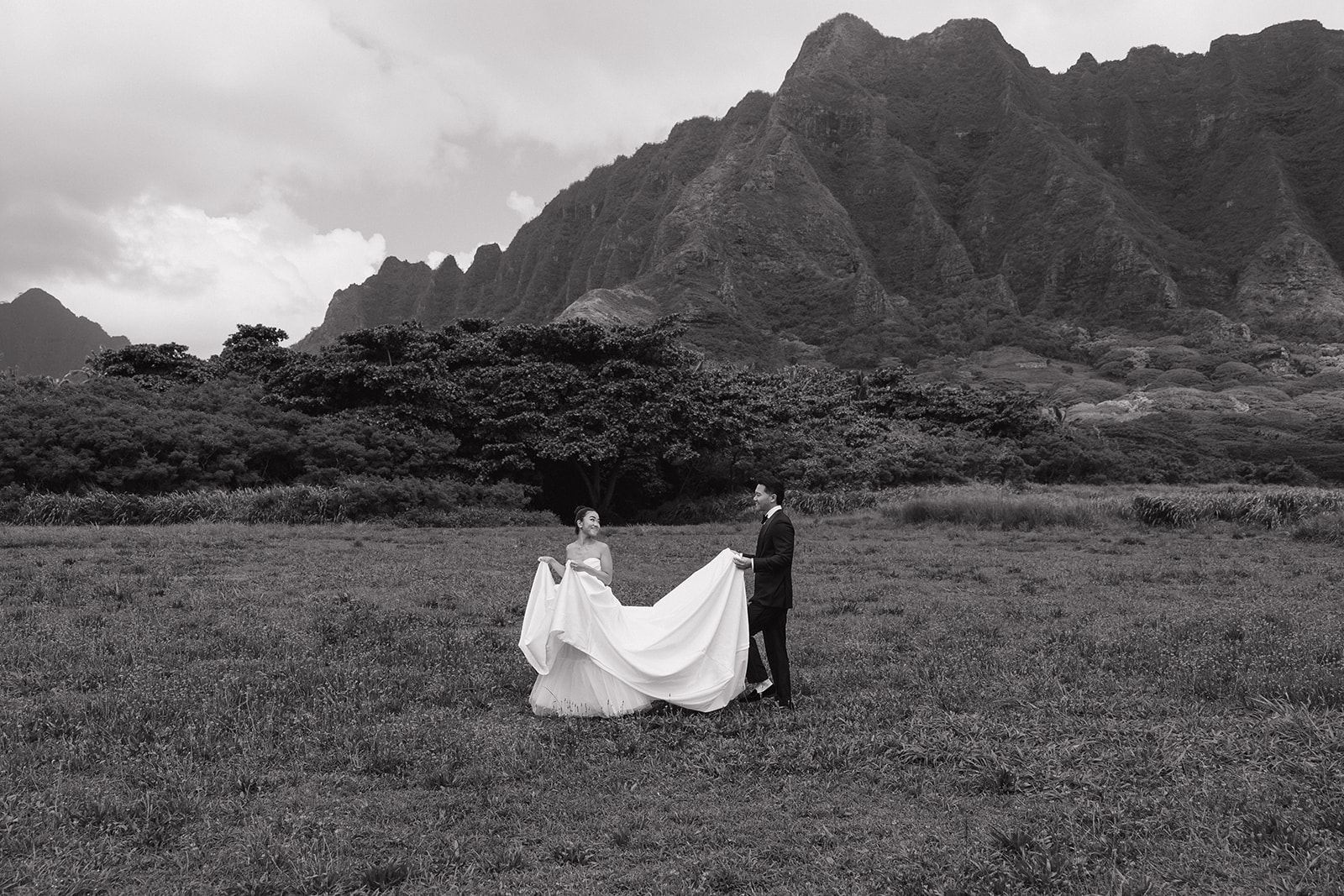 Black and white wide-angle photo of bride and groom playfully spreading her ball gown train across an open field at their Kualoa Ranch Wedding, with the full dramatic span of the Ko'olau mountain range filling the entire background