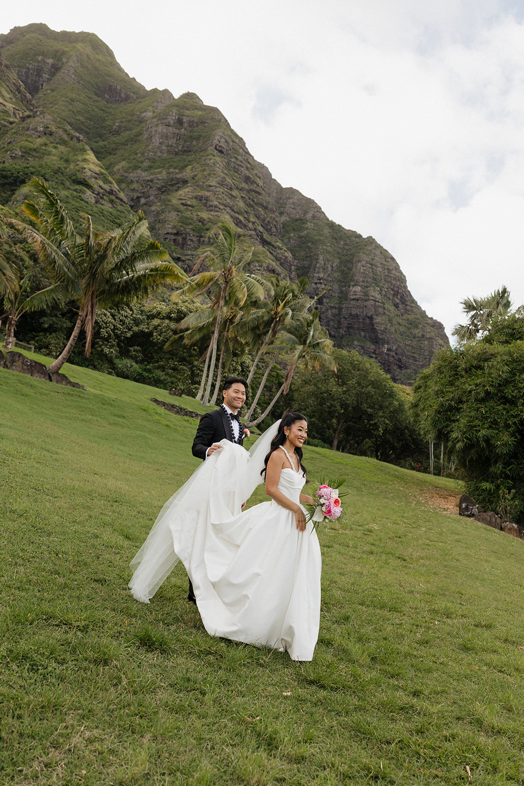 Bride walking up a grassy slope holding her tropical bouquet while her groom carries her flowing veil behind her at their Kualoa Ranch Wedding, with towering jagged Ko'olau cliffs and swaying palms rising dramatically overhead