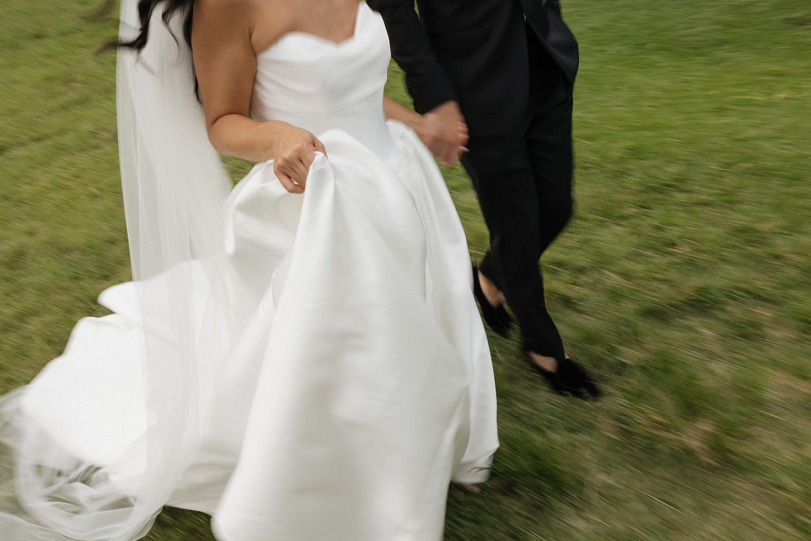 Close-up action shot of bride gathering her white satin ball gown train while running hand-in-hand with her groom across a grassy Hawaiian field