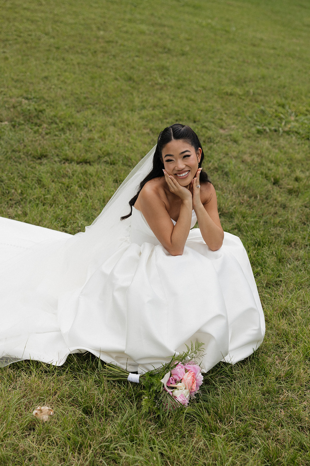 Radiant bride sitting on the lawn and propping her chin in her hands with a beaming smile, her full white ball gown and cathedral veil pooling around her alongside her pink peony bouquet