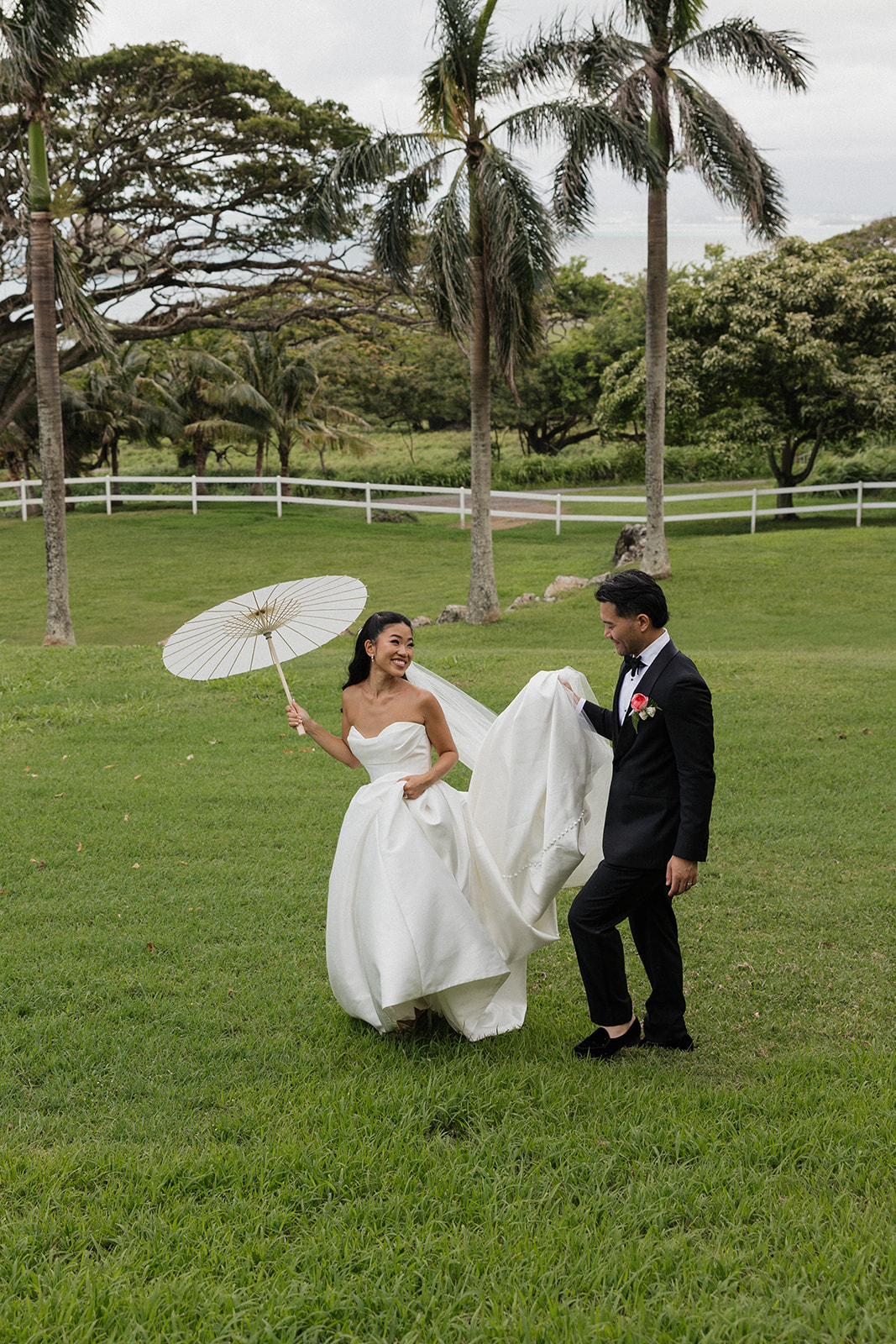 Smiling bride holding a white paper parasol while her groom carries the cathedral train of her strapless ball gown across the grass at their Kualoa Ranch Wedding, with tall royal palms and a white picket fence in the background