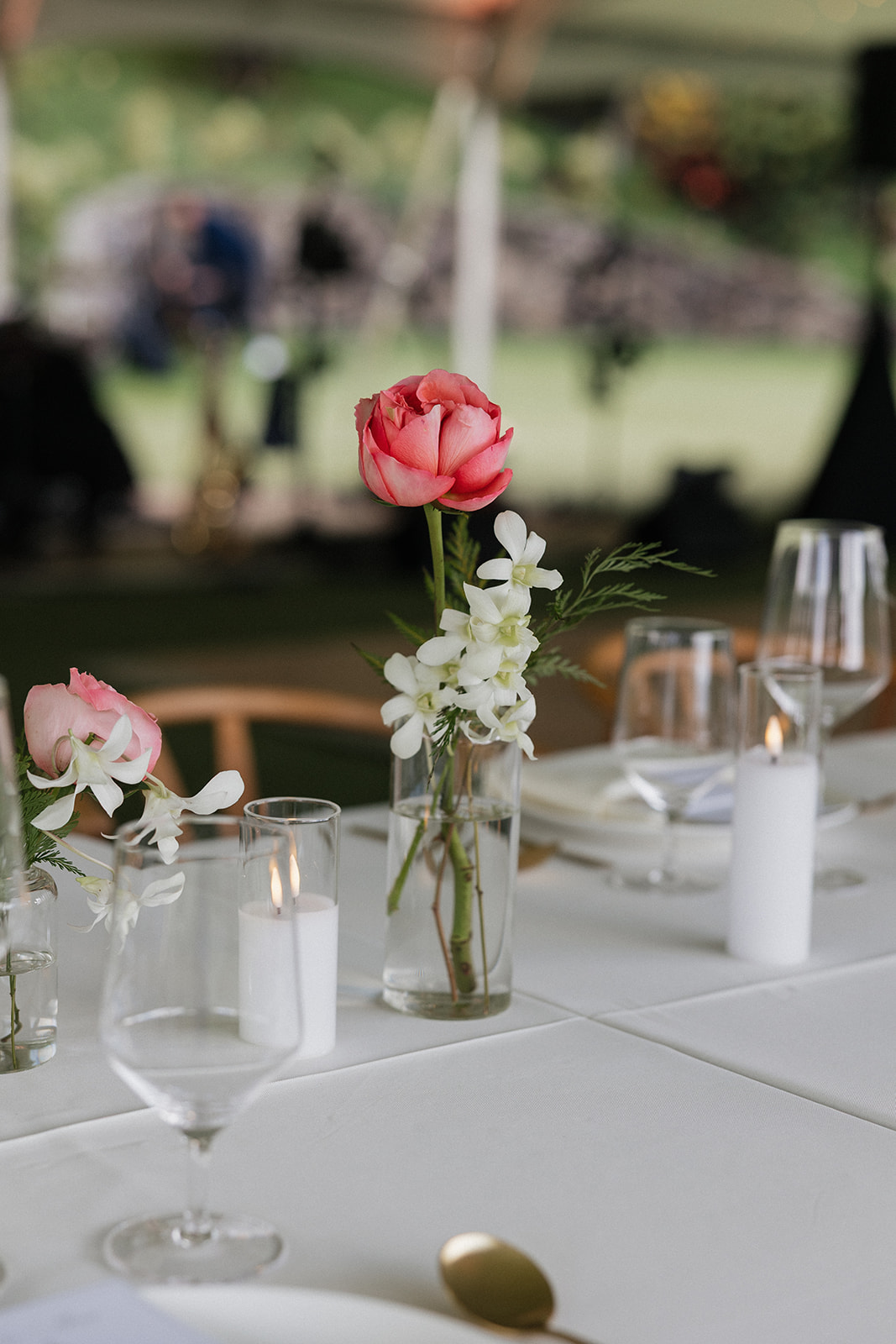 Close-up of a minimalist Kualoa Ranch Wedding reception centerpiece — a single coral peony and white stock stems in a glass bud vase alongside a lit pillar candle on a white linen tablecloth