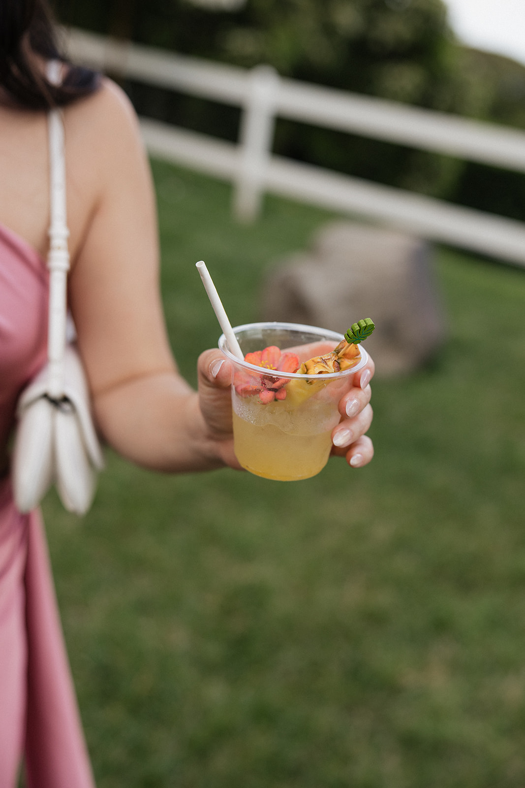 Guest in a blush dress holding a tropical signature cocktail garnished with a fresh hibiscus flower, pineapple wedge, and decorative leaf pick at an outdoor Hawaiian wedding cocktail hour