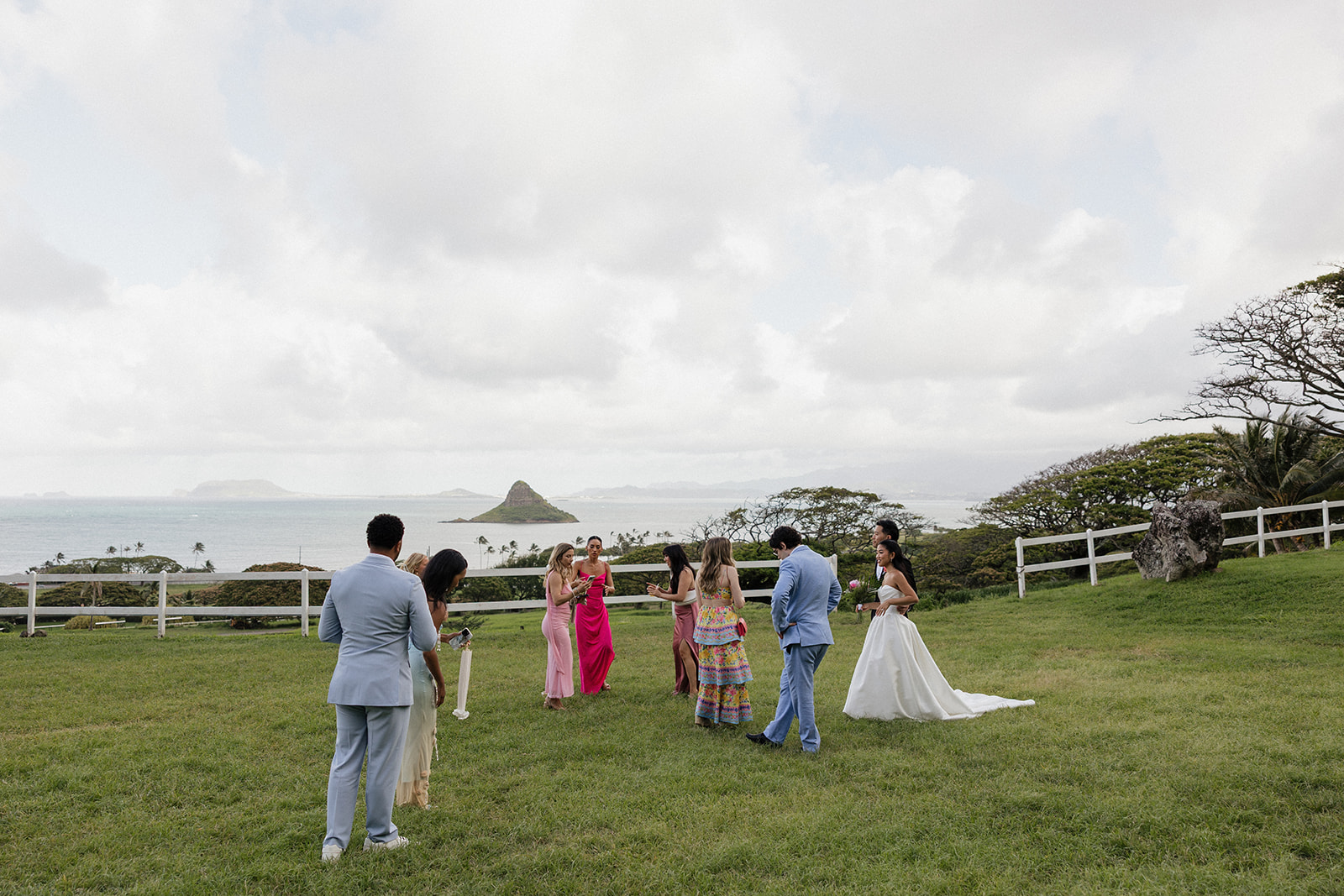 Guests and the bride mingling on the open lawn at a Kualoa Ranch Wedding cocktail hour, with Mokoli'i Island and the windswept coast of Oahu's Kaneohe Bay stretching out behind a white picket fence