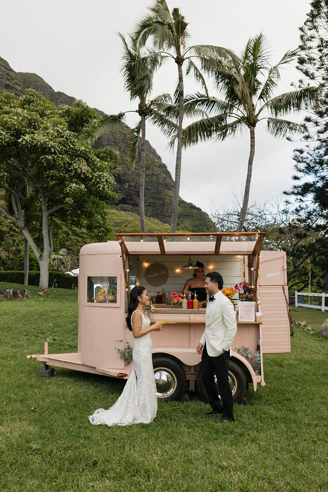 Bride and groom laughing together beside a vintage blush pink cocktail trailer serving colorful drinks, with tall palm trees and the dramatic Ko'olau mountains rising behind them