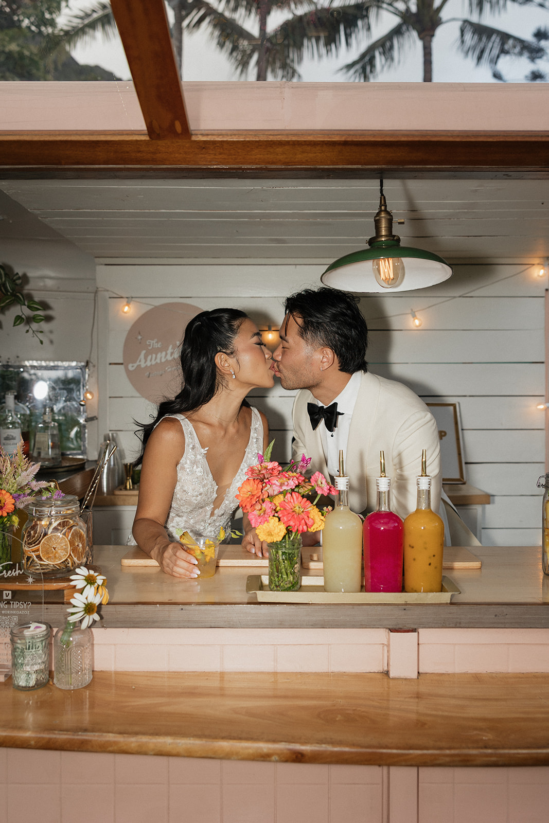 Bride and groom stealing a kiss across the counter of a charming pink vintage bar trailer at their Kualoa Ranch Wedding cocktail hour, surrounded by bright tropical flowers and colorful drink syrups