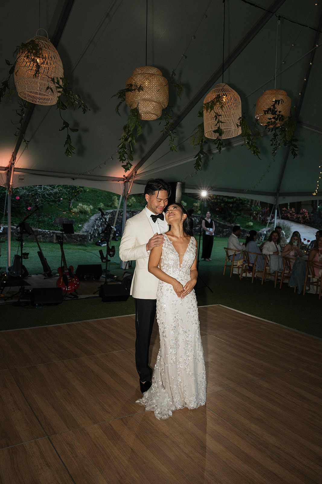 Bride and groom sharing their first dance under woven rattan pendant lanterns draped with greenery inside a tented Kualoa Ranch Wedding reception, with a live band and warm bistro lighting in the background