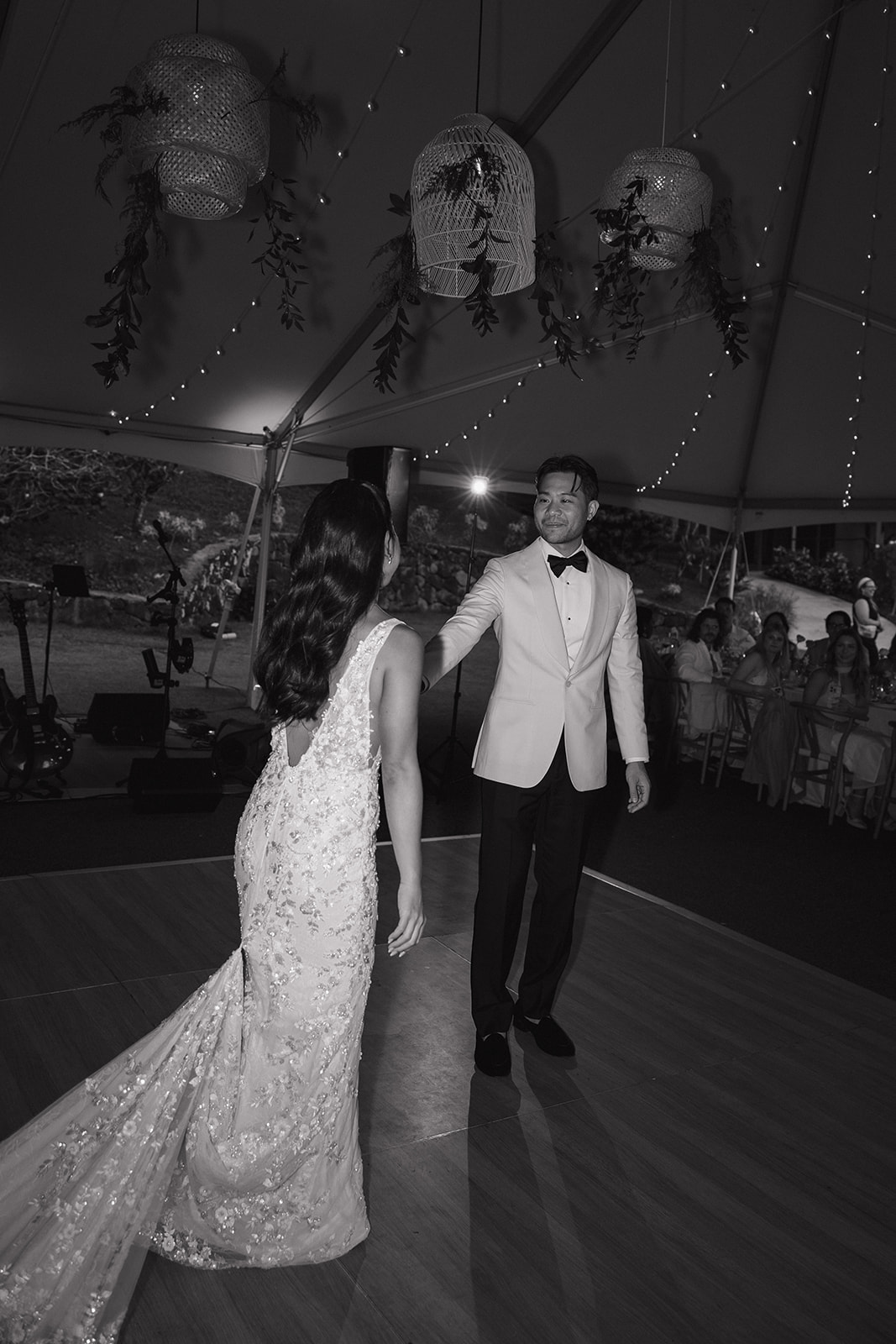 Black and white first dance photo at a Kualoa Ranch Wedding reception — groom in a white dinner jacket reaching out to his bride on the wooden dance floor under glowing rattan lanterns and fairy lights, with seated guests watching in the background