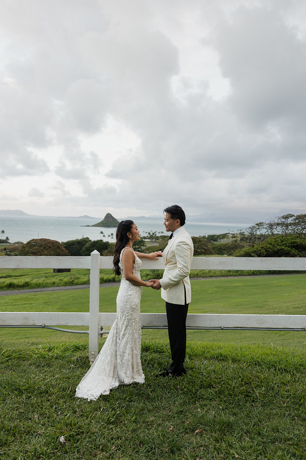 Bride in a beaded lace gown and groom in a white dinner jacket holding hands and facing each other at their Kualoa Ranch Wedding, with the iconic Mokoli'i Island rising from the ocean behind a white fence