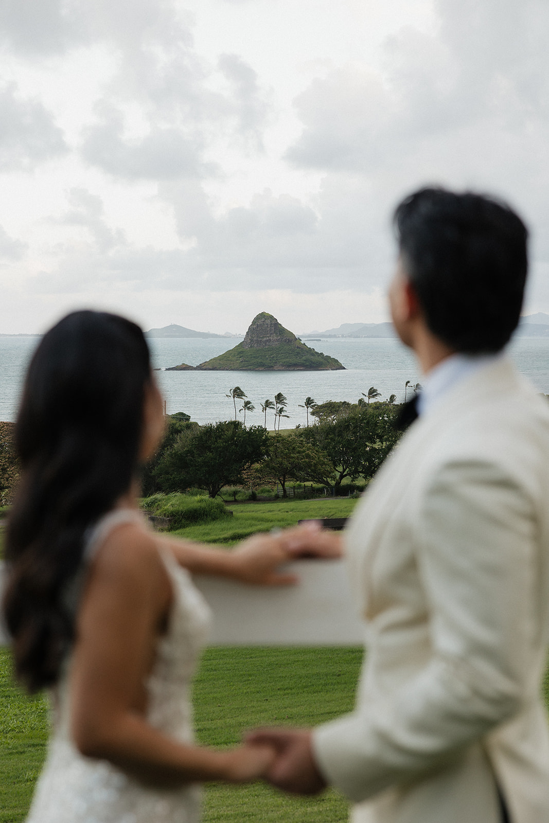 Couple standing together in soft focus on a hillside, gazing toward Mokoli'i Island rising from the turquoise waters of Kaneohe Bay on Oahu's windward shore