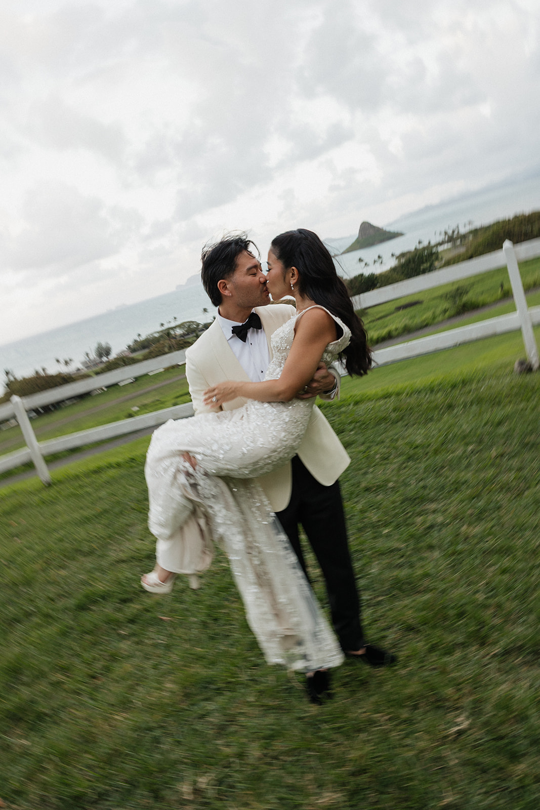 Groom lifting bride off her feet for a spinning kiss on the green lawn with Mokoli'i Island and the turquoise waters of Kaneohe Bay visible behind a white fence in the background