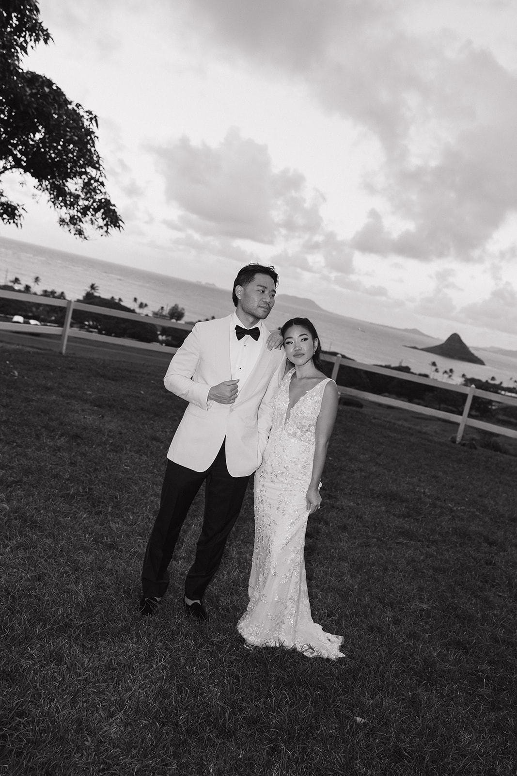 Black and white evening portrait of bride and groom posing confidently on a grassy hillside at their Kualoa Ranch Wedding, with Mokoli'i Island silhouetted against a dramatic cloudy sky over the ocean