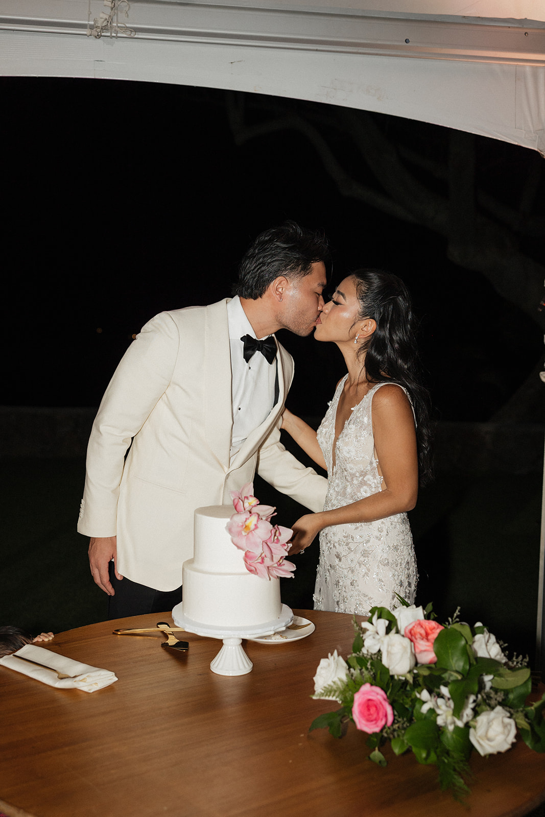 Bride and groom sharing a kiss over their two-tier white wedding cake decorated with pink orchids, with a gold cake server on the table and a garden rose centerpiece at their Hawaiian reception
