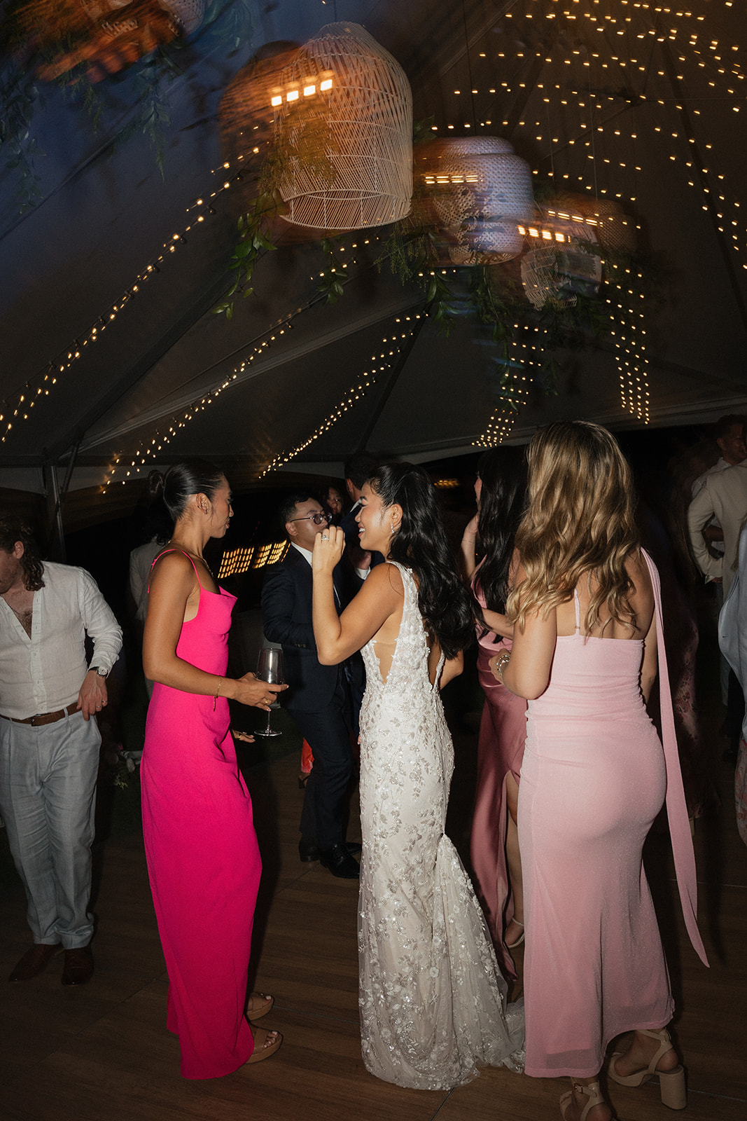 Bride in a beaded lace gown dancing and laughing on the floor with guests under rattan pendant lanterns and thousands of twinkling fairy lights strung across the ceiling of the reception tent