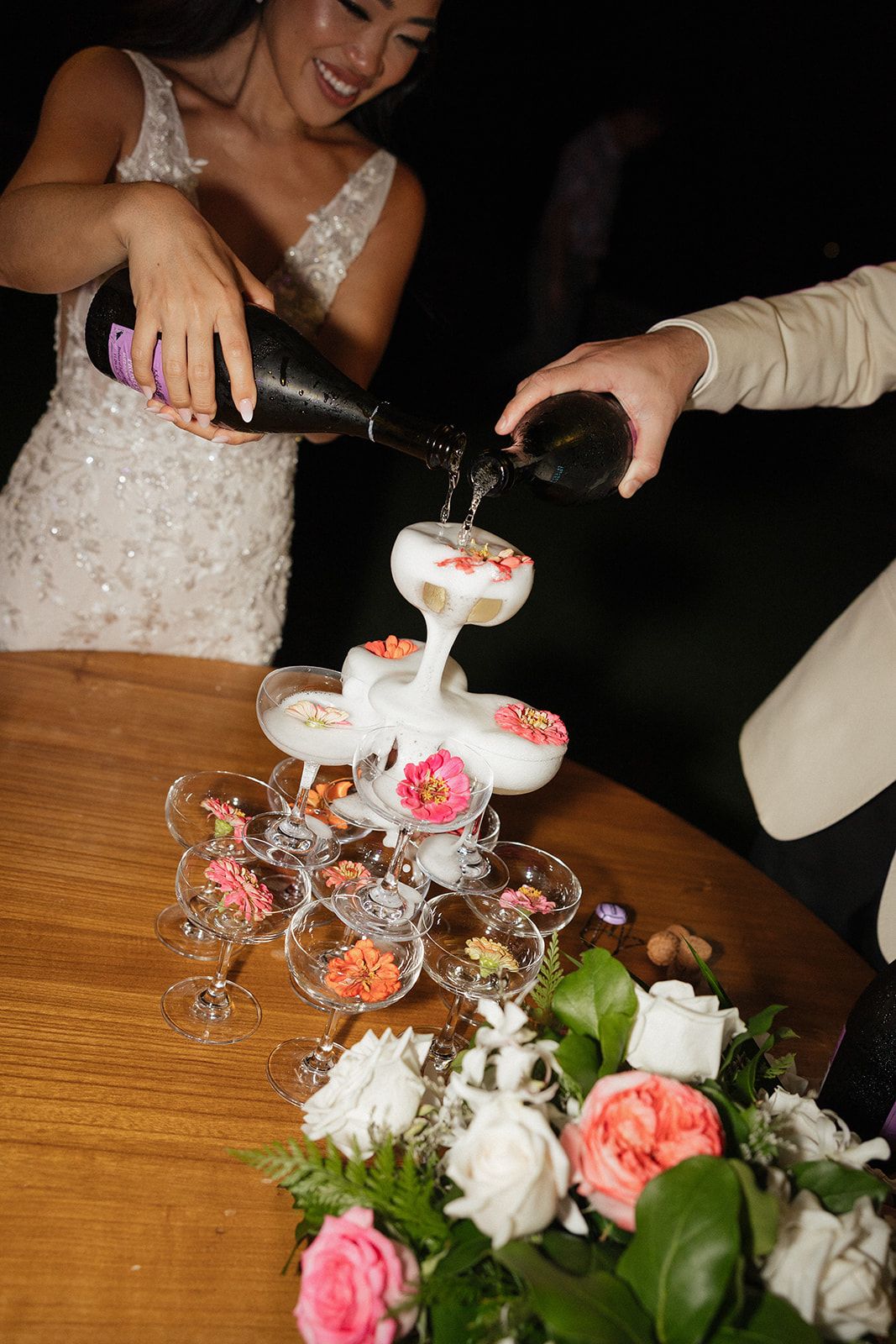 Bride and groom pouring champagne together over a flower-adorned coupe tower at their Kualoa Ranch Wedding reception, with a lush floral centerpiece of pink and white roses on the wooden table below