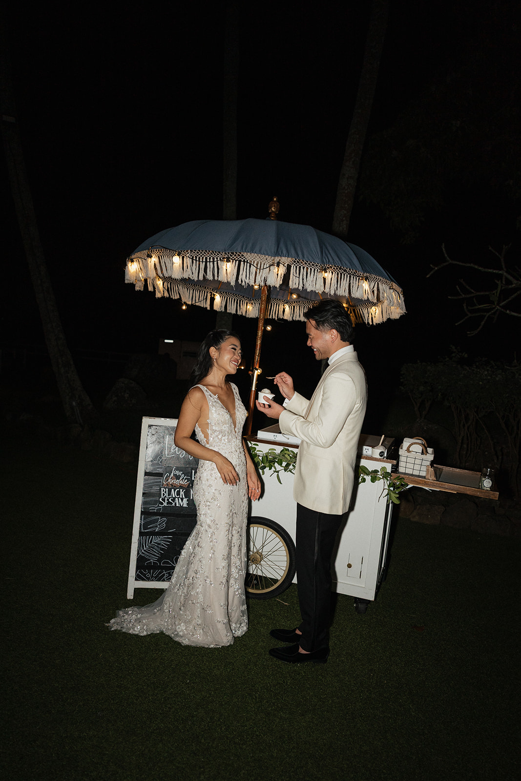 Bride and groom laughing together at a vintage white ice cream cart with a macramé-fringed blue umbrella strung with fairy lights during their outdoor Hawaiian wedding reception at night