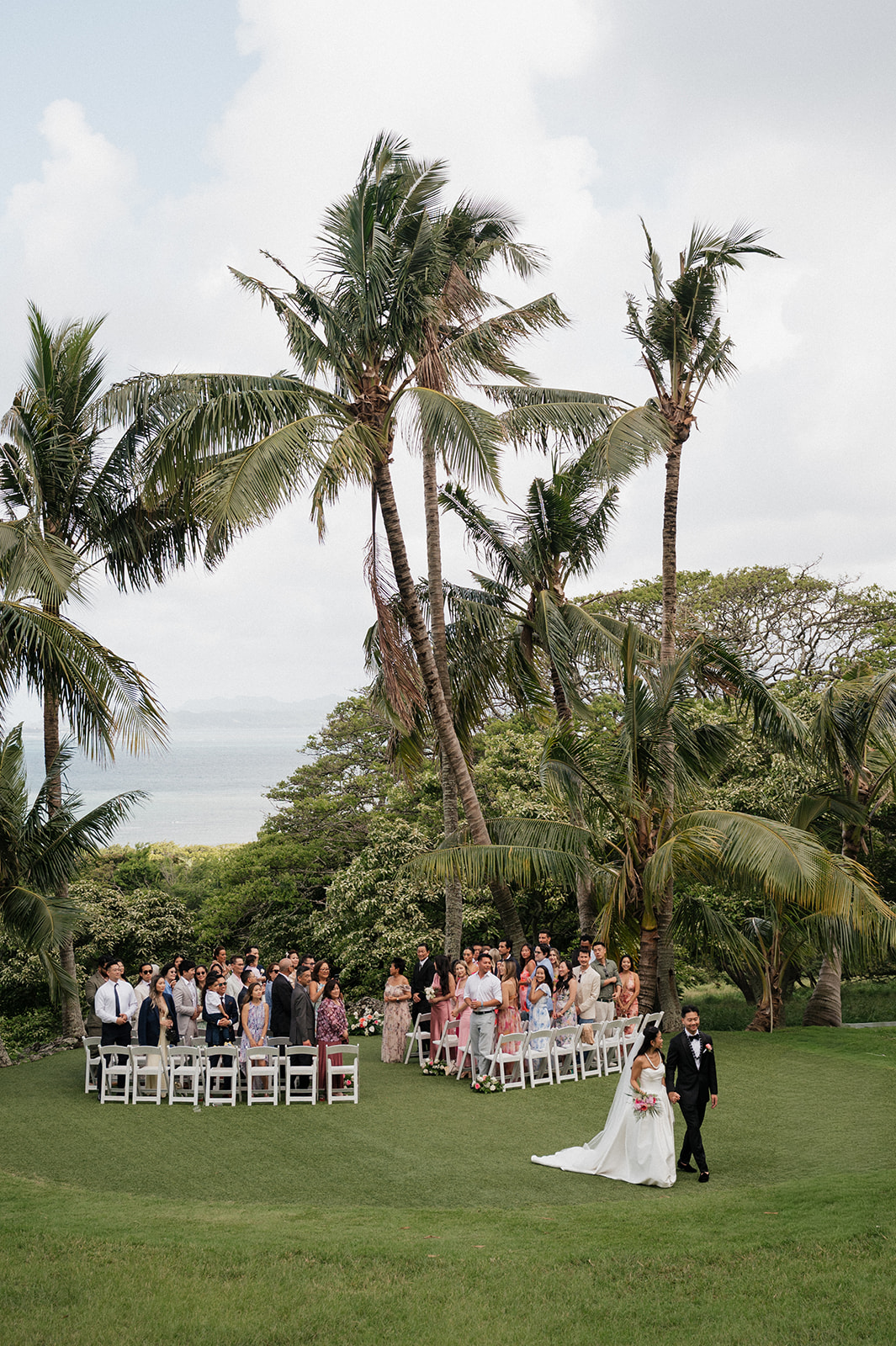 Wide-angle shot of the outdoor wedding ceremony at Kualoa Ranch Wedding venue, with guests seated in white chairs among towering coconut palms and the Pacific Ocean visible in the distance