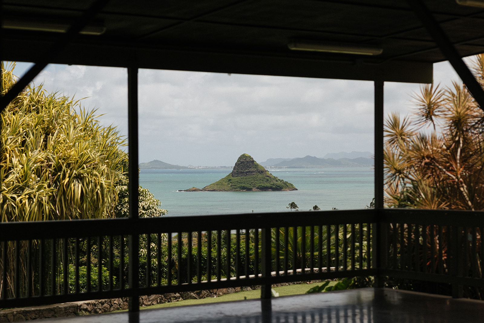 View of Mokoli'i Island (Chinaman's Hat) framed through a dark wooden pavilion railing, with turquoise water and tropical vegetation on Oahu's windward coast