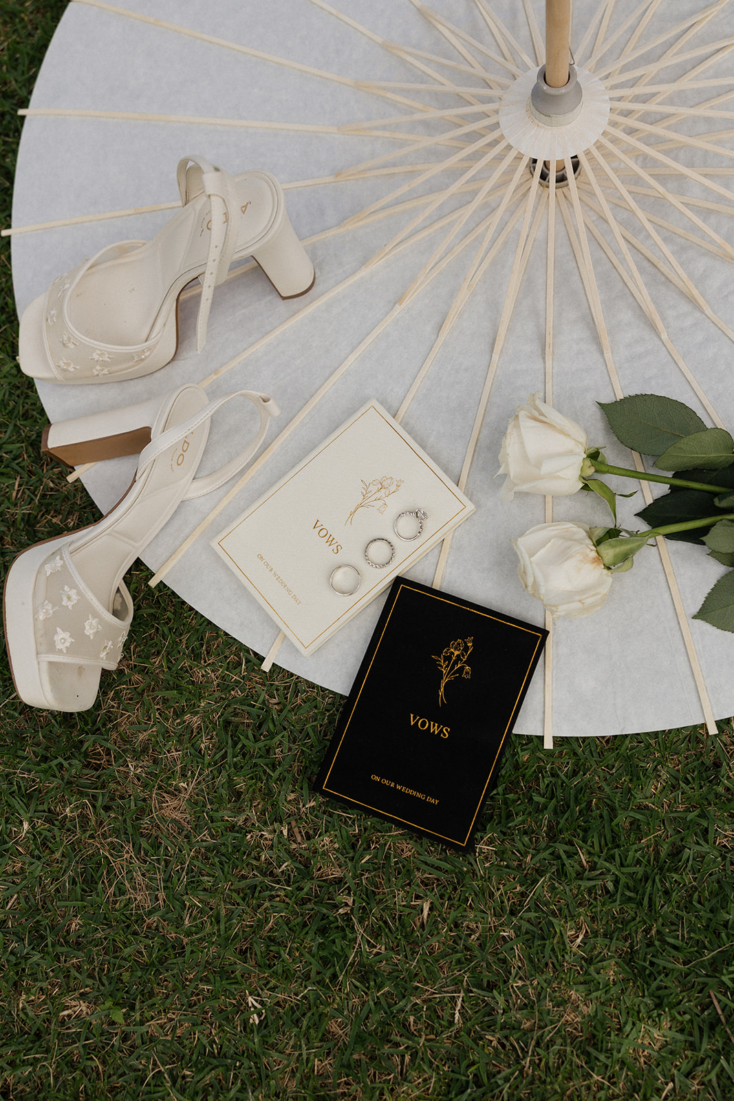 Flat lay of Kualoa Ranch Wedding details, white floral heeled bridal shoes, coordinating black and ivory vow books with gold foil lettering, wedding rings, and white roses on a parasol
