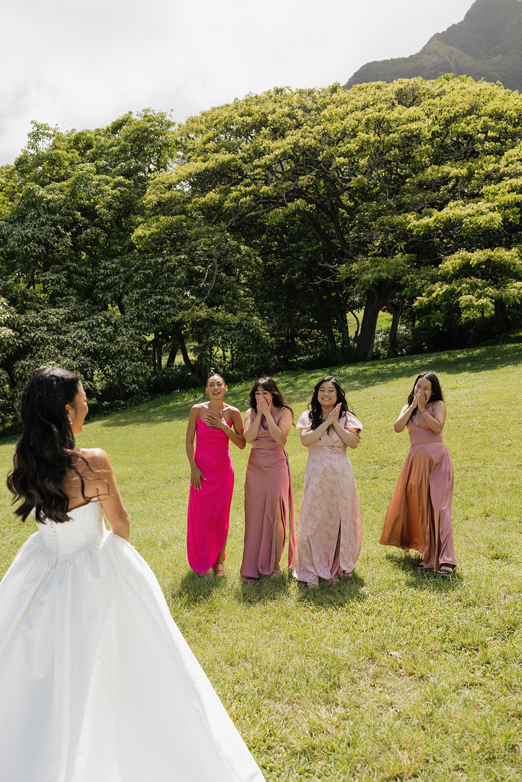 Bridesmaids reacting with delight and tearful smiles during a first look reveal at a Kualoa Ranch Wedding, wearing coordinating shades of hot pink, dusty rose, and terracotta