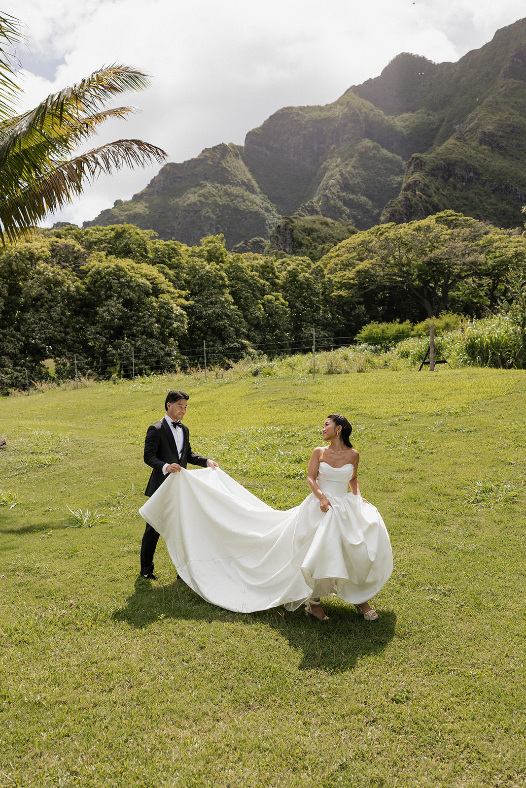 Groom in a classic black tuxedo fluffing and spreading the cathedral train of his bride's strapless ball gown across the bright green grass, with sunlight breaking through clouds over the Ko'olau mountain peaks