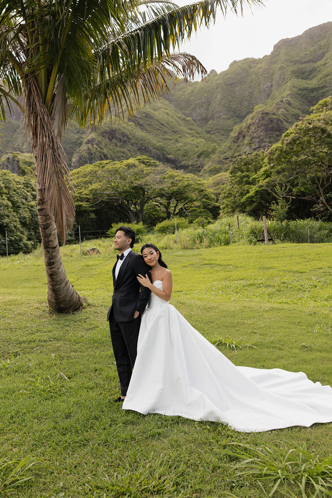 Bride and groom posing under a palm tree at their Kualoa Ranch Wedding, with dramatic Ko'olau mountain cliffs rising behind them on Oahu